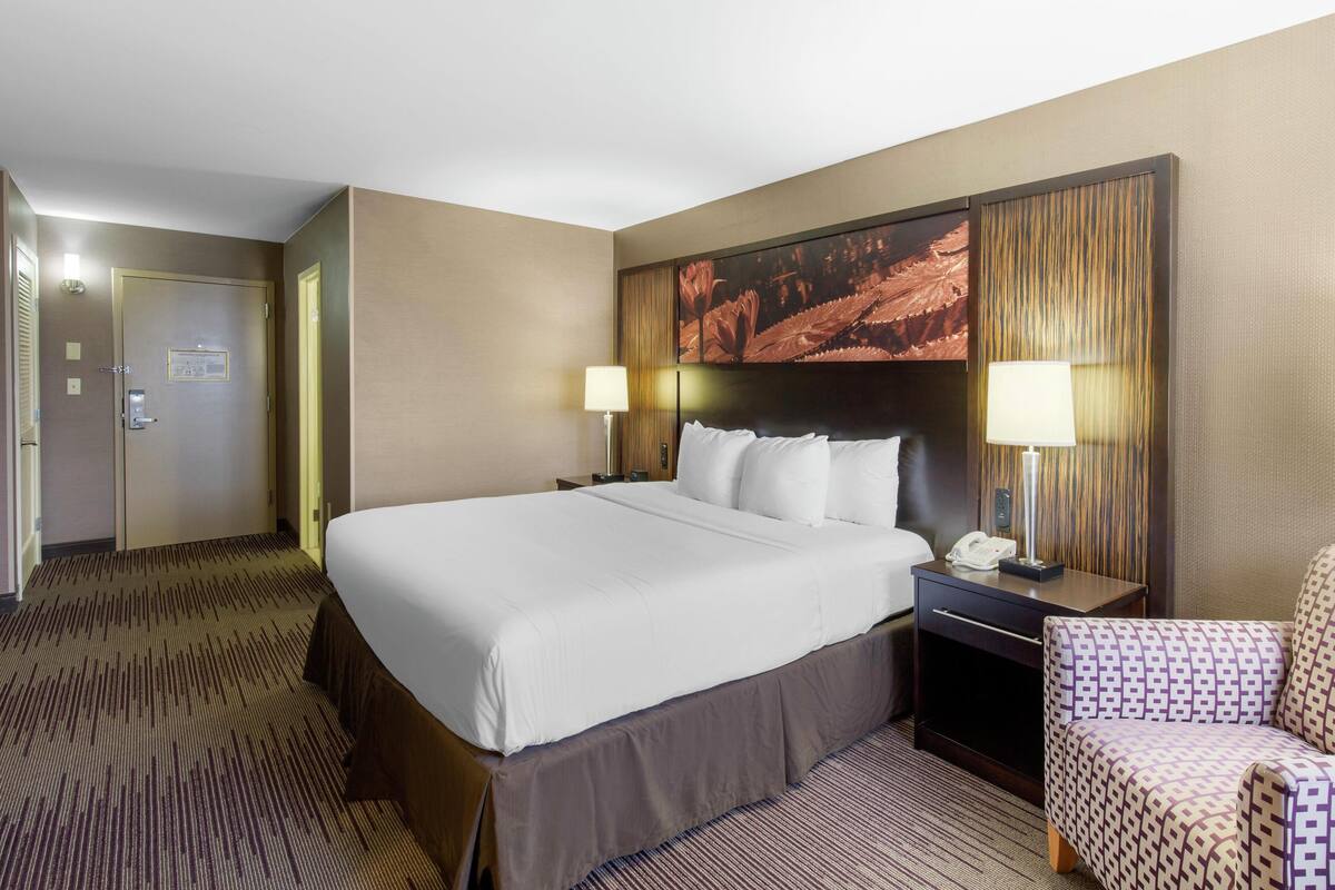 A hotel room with a large bed, two bedside lamps, a nightstand, a chair, and a large wall painting.