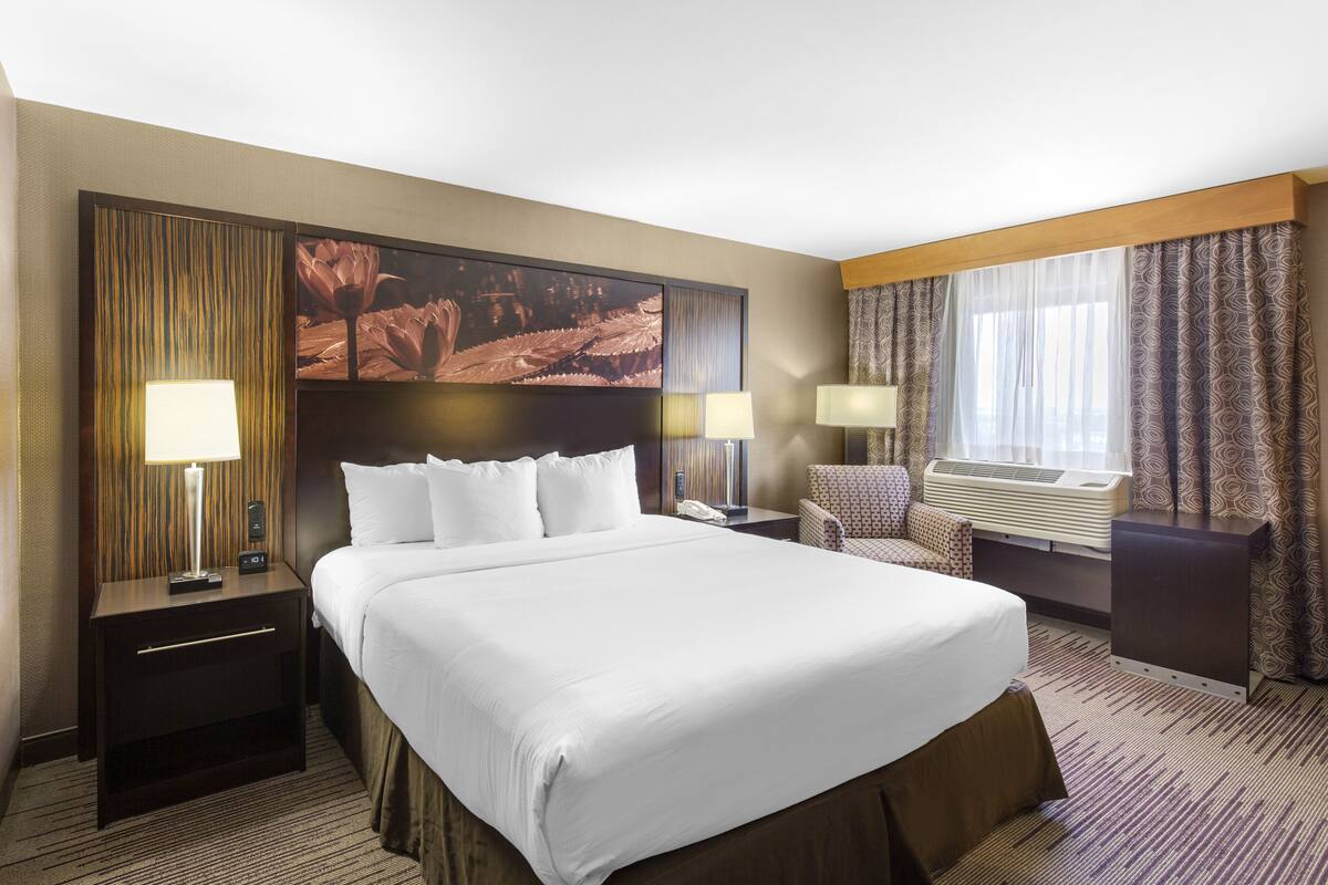 A hotel room with a large bed, two bedside tables with lamps, a chair, a large painting on the wall, and a window with curtains.