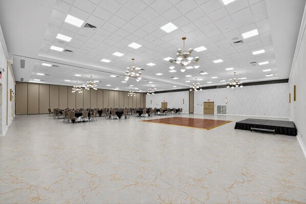 A spacious conference room with a central dance floor, multiple tables with chairs, and a modern chandelier.