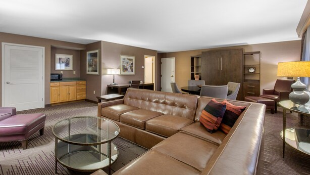 A modern living room with a brown leather sofa, a glass coffee table, and a dining area with a wooden cabinet and a small table. - DoubleTree by Hilton Downtown Wilmington - Legal District (Wilmington)