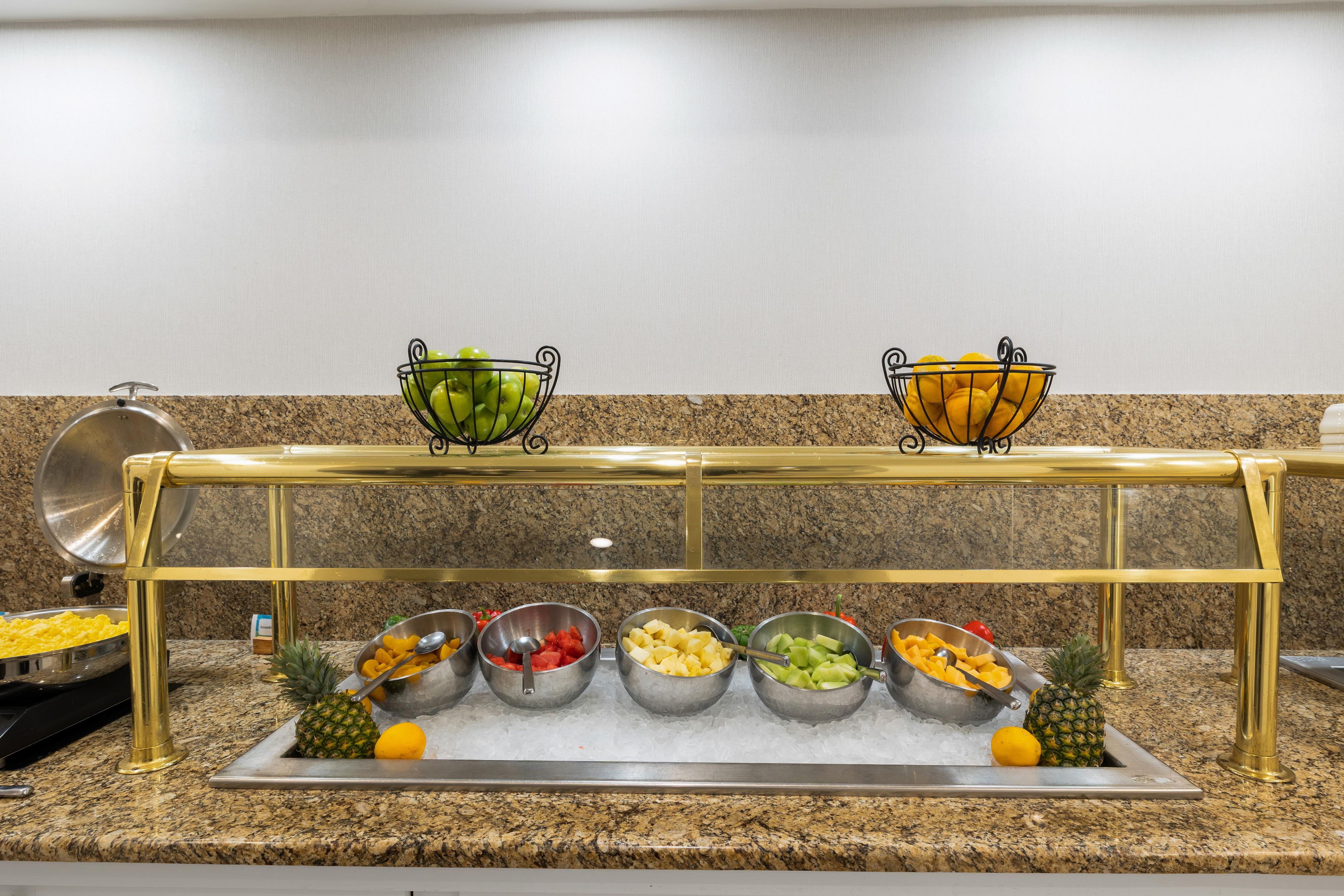 A buffet setup with various fruits and vegetables in metal bowls, a pineapple, and a metal tray with a spoon.