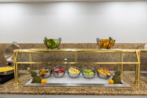 A buffet setup with various fruits and vegetables in metal bowls, a pineapple, and a metal tray with a spoon.