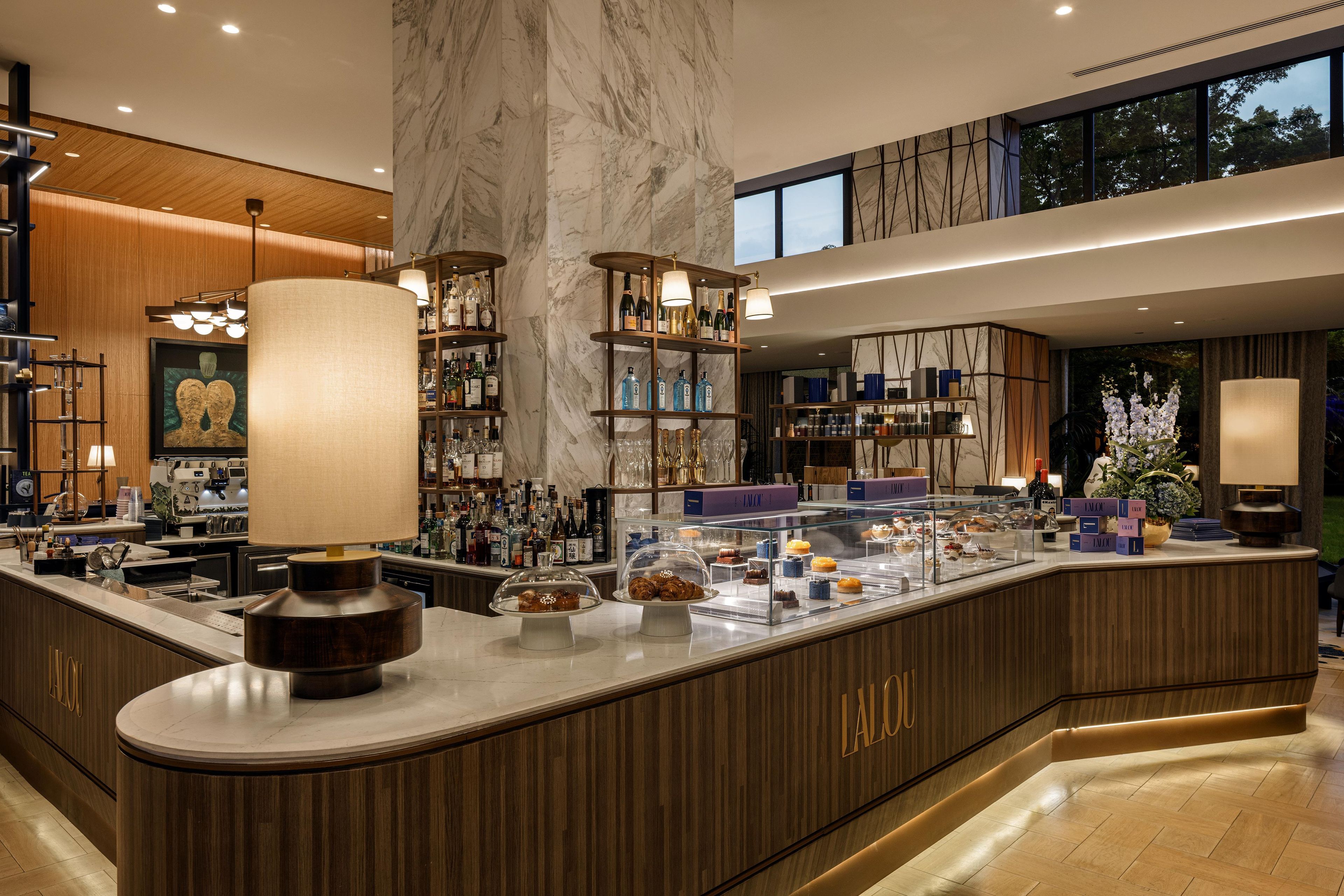 Un espace bar moderne avec un sol en marbre, des étagères en bois remplies de bouteilles et une vitrine où sont exposées des pâtisseries.
