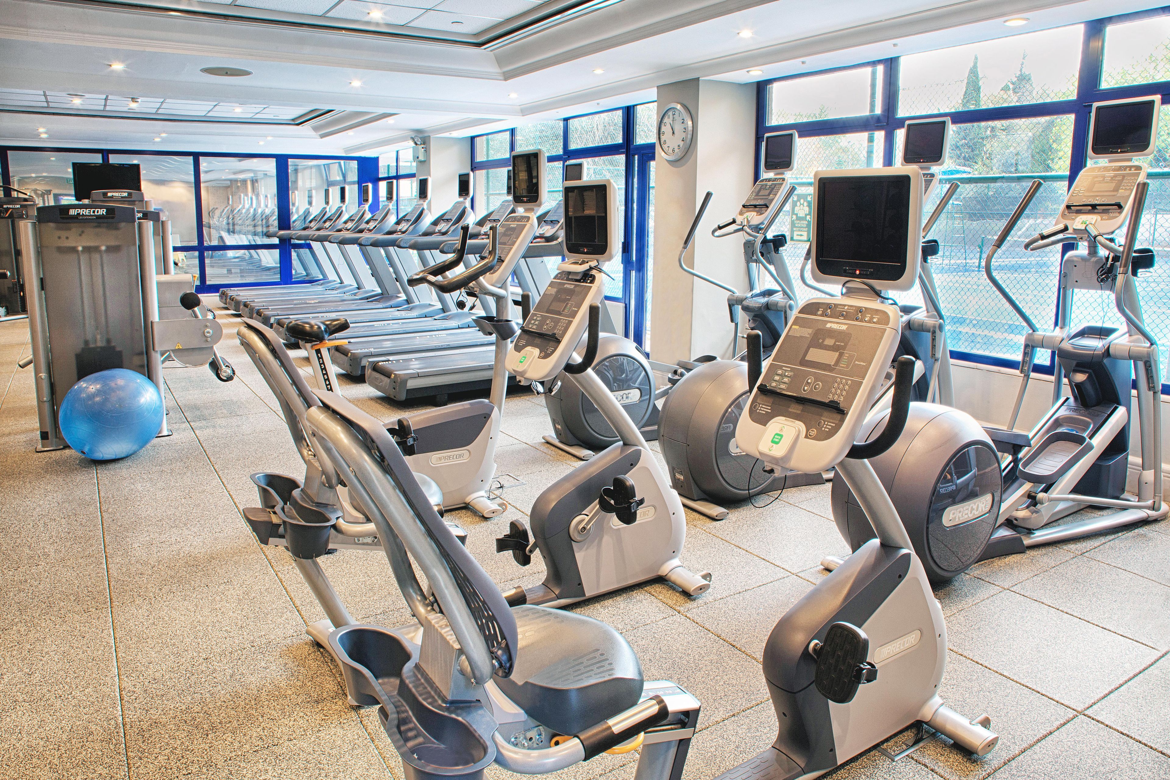 A gym with multiple exercise machines, a large window with a view, and a clock on the wall.
