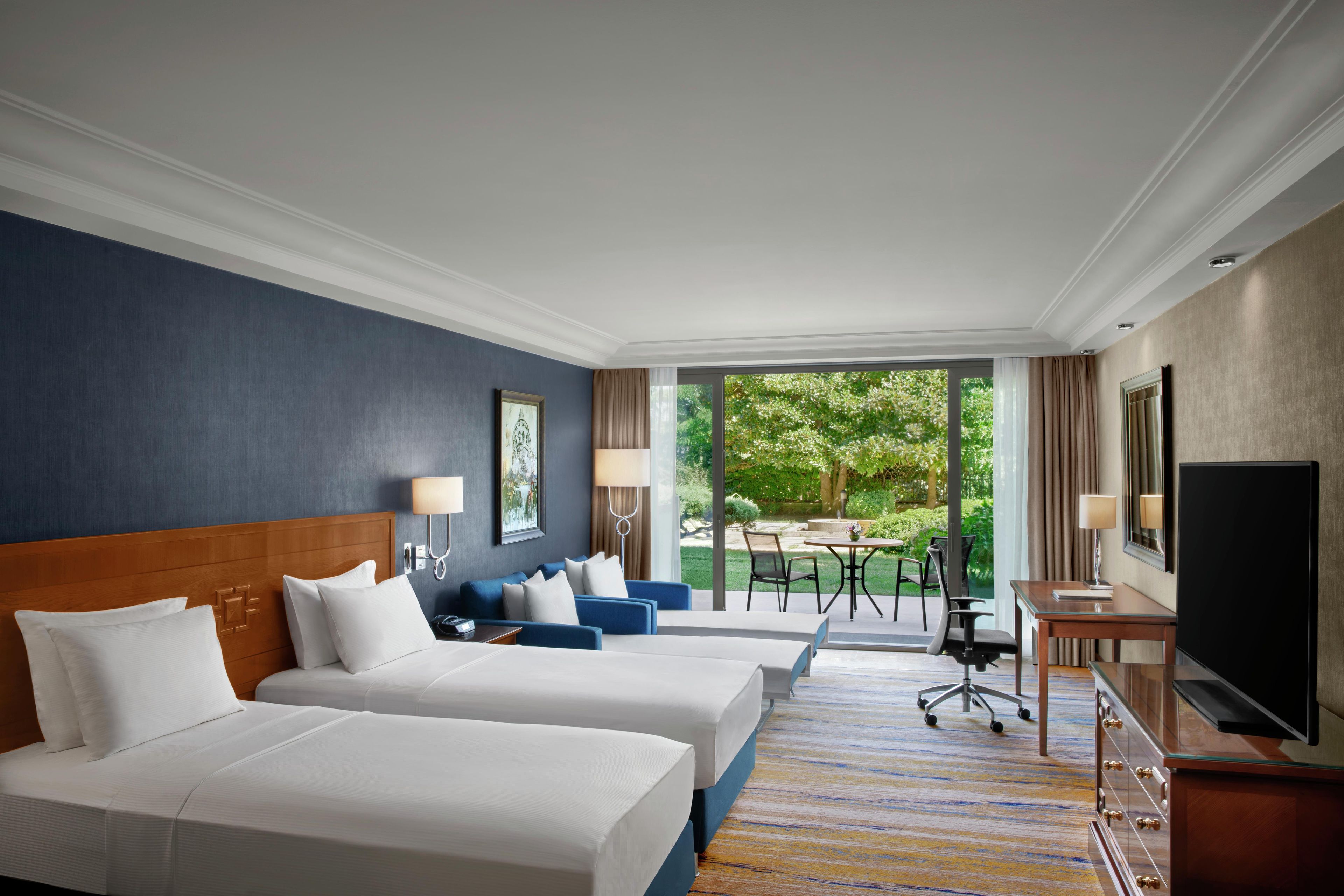 Ein Hotelzimmer mit zwei Betten, einem Schreibtisch, einem Fernseher und einem Balkon mit Blick auf Grünflächen.