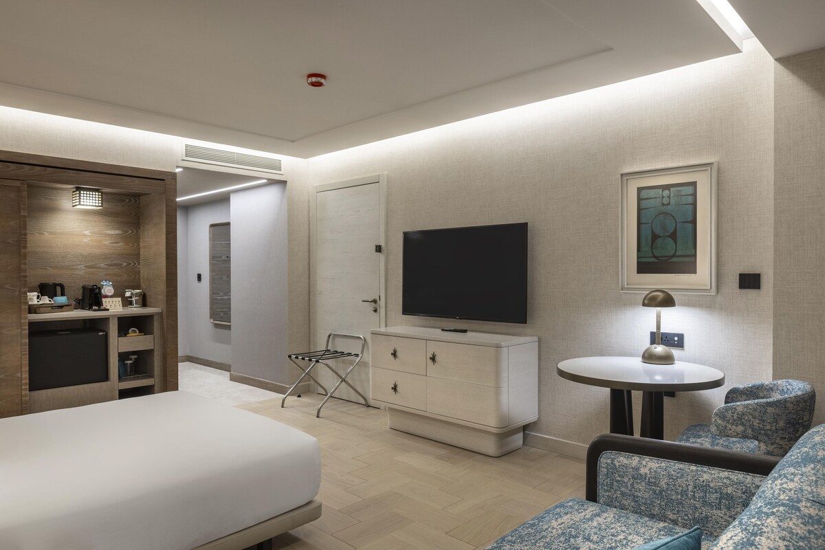 Una habitación de hotel moderna con cama, zona de estar, televisión, mesita y kitchenette.