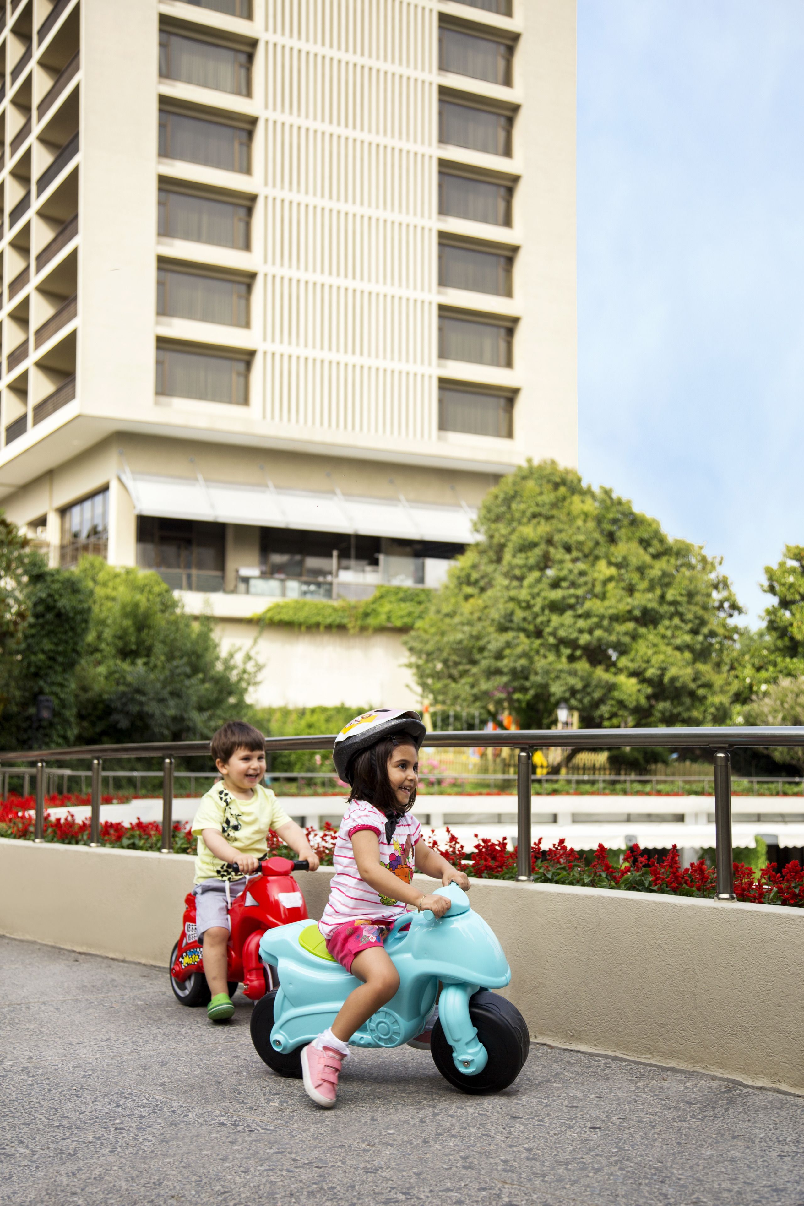 Two children riding scooters on a paved path with a building and trees in the background.