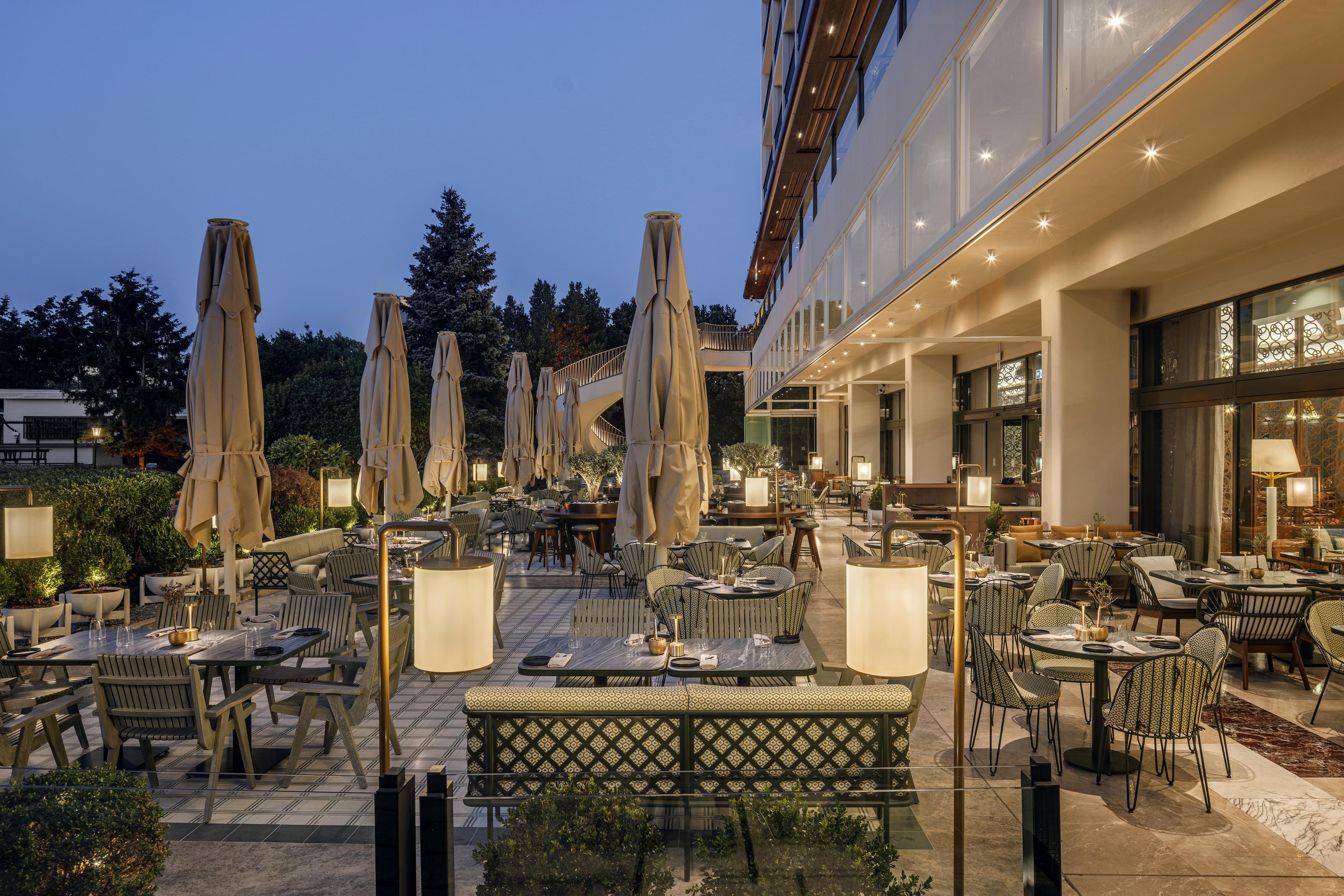 An outdoor dining area with tables and chairs, umbrellas, and a building with large windows in the background.
