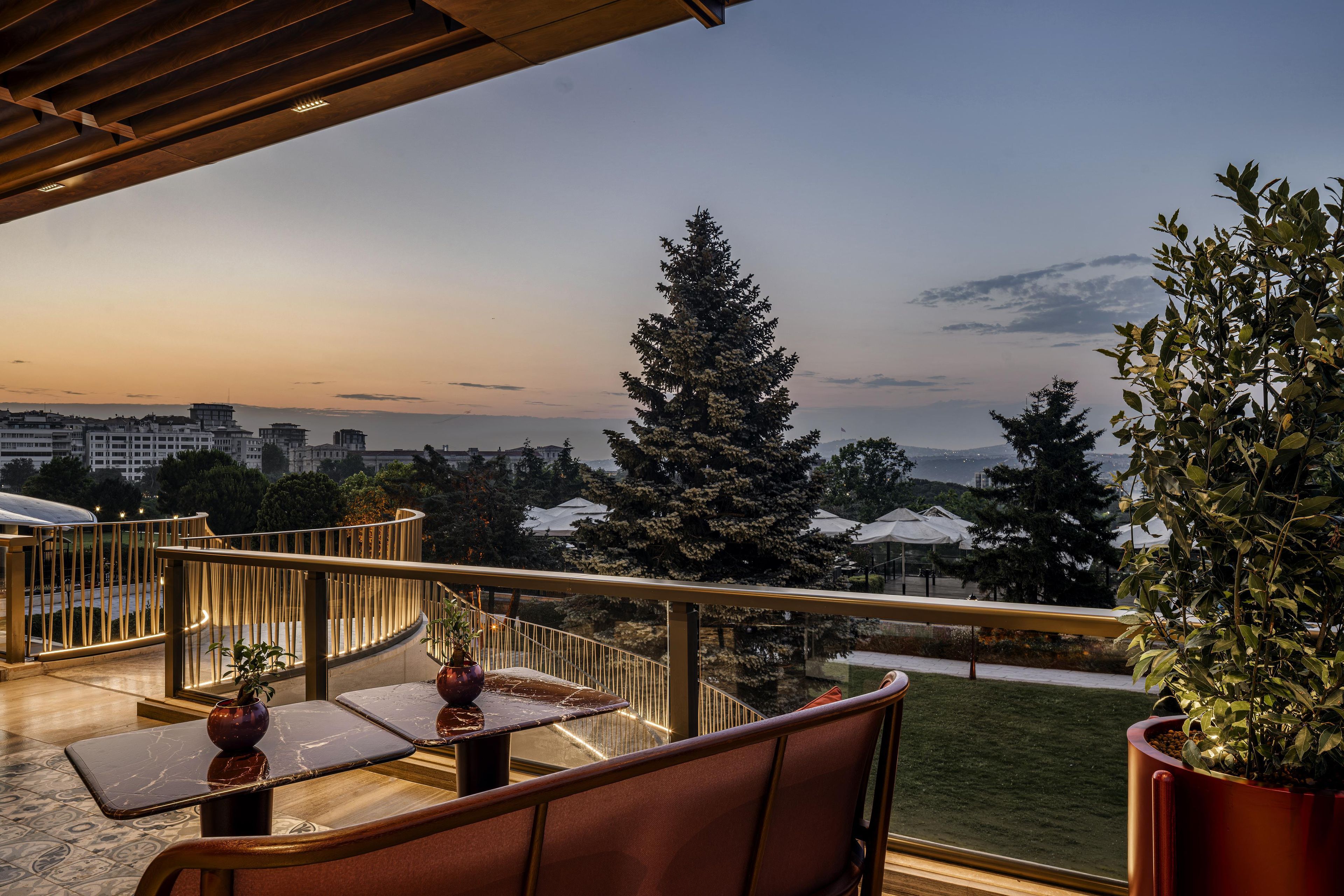 A balcony with a view of a cityscape at dusk, featuring a railing, seating area, and potted plants.
