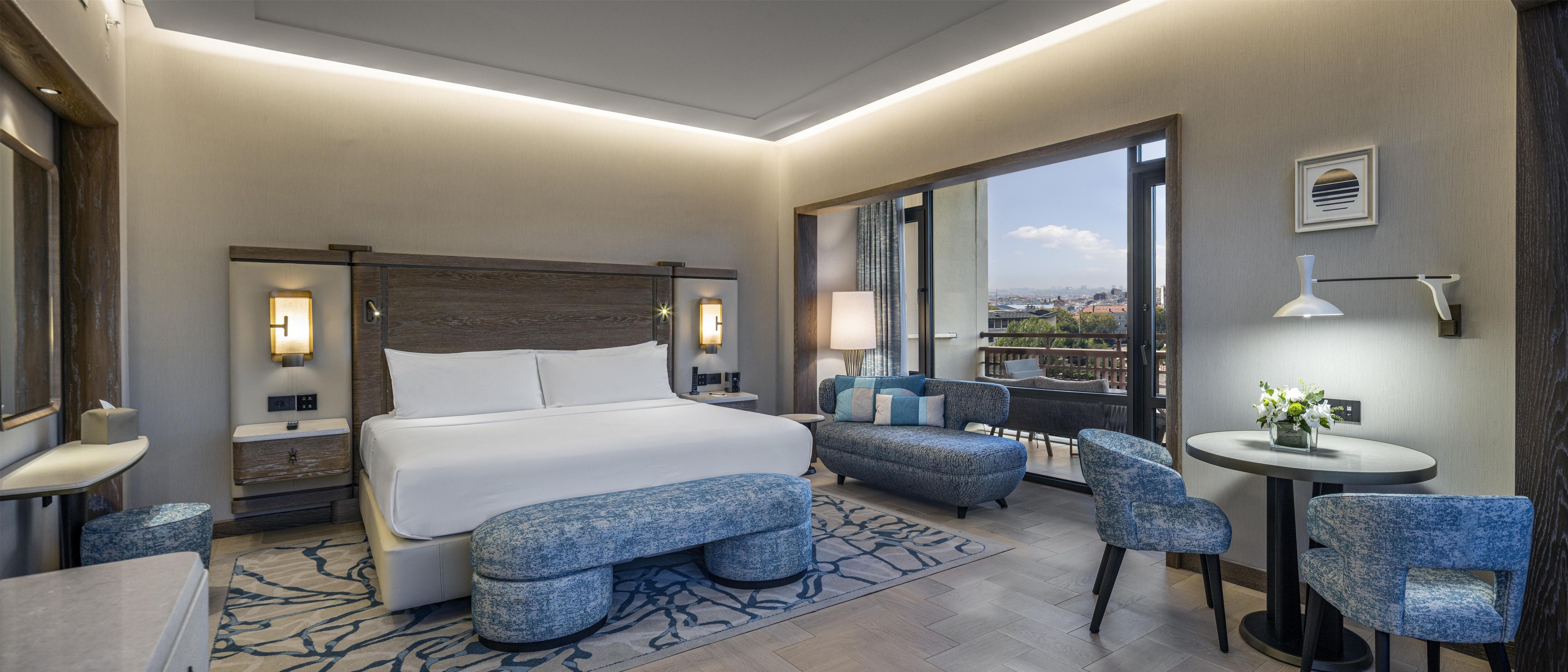 A hotel room with a large bed, a seating area with chairs, a small table, and a view through a large window.