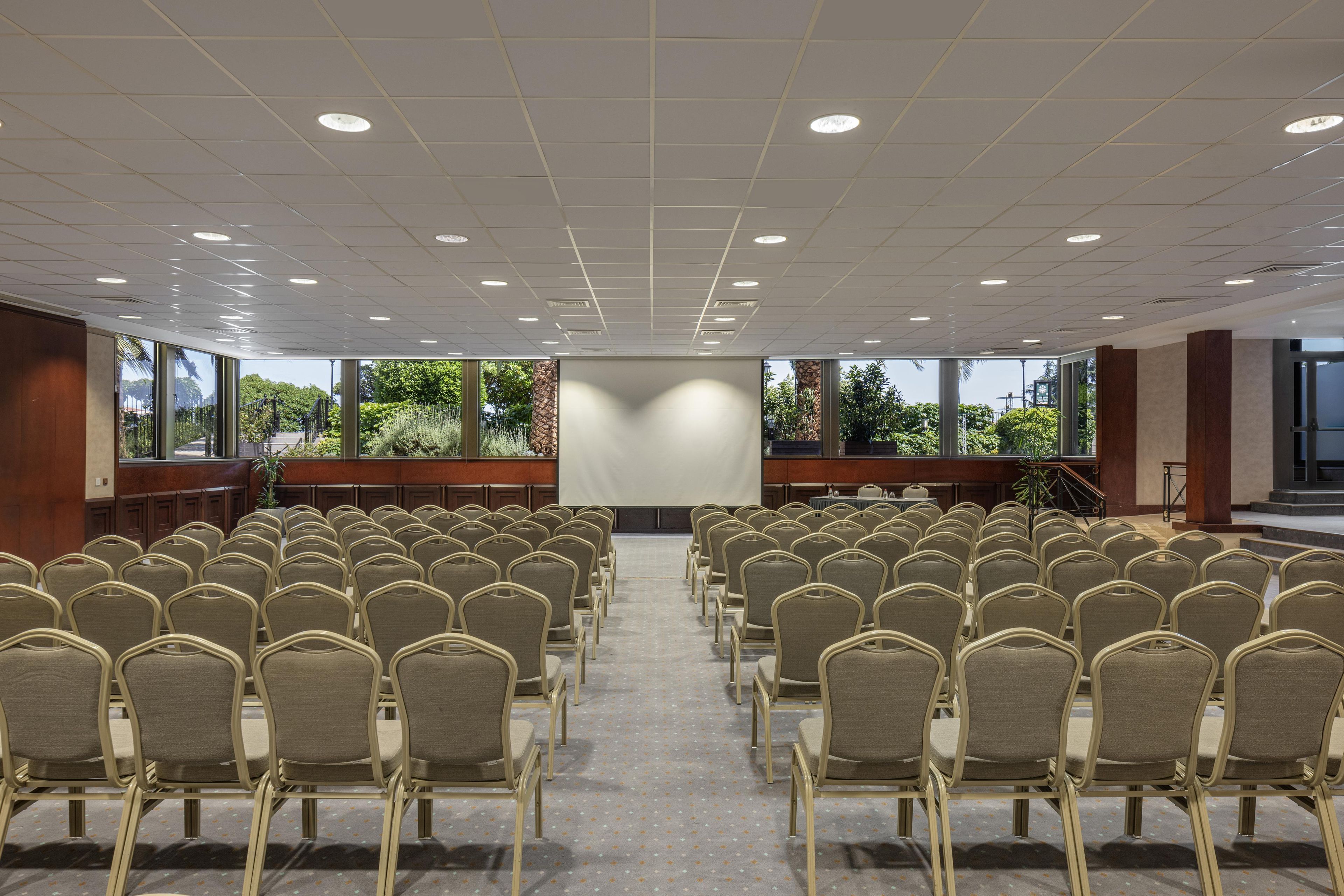 A conference room with rows of chairs facing a projection screen, large windows with a view of greenery, and a stage with a podium.