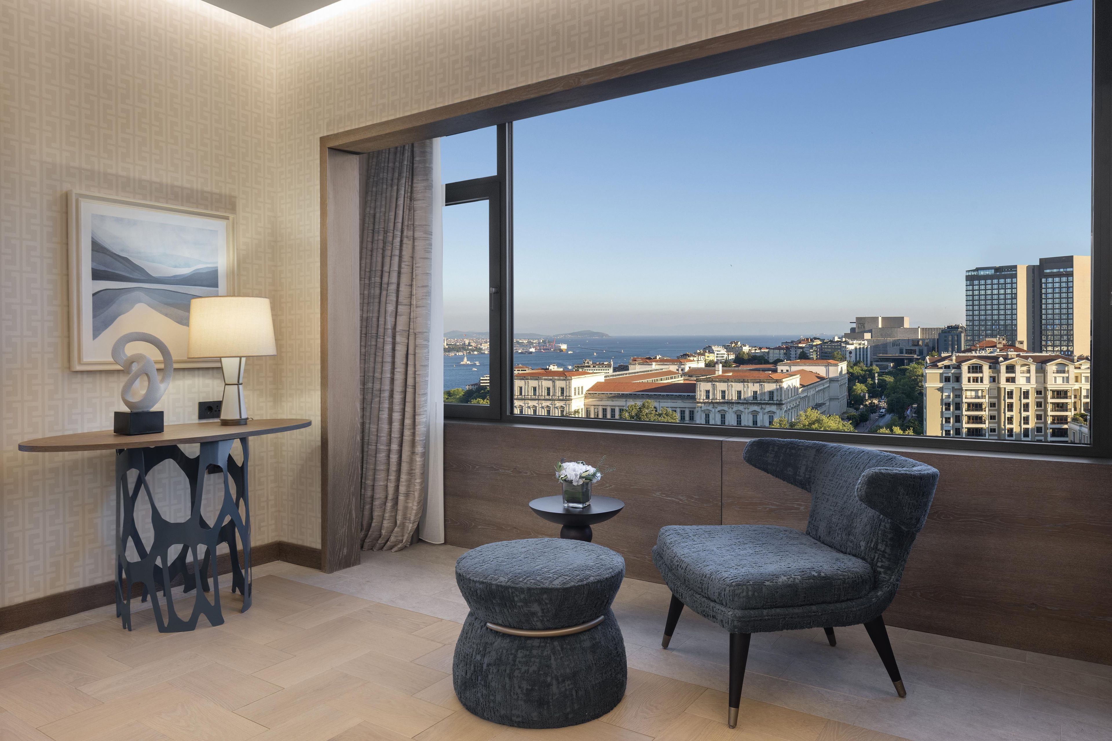 A hotel room with a large window, a desk, a chair, a small table, and a decorative lamp.