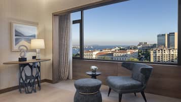A hotel room with a large window, a desk, a chair, a small table, and a decorative lamp.
