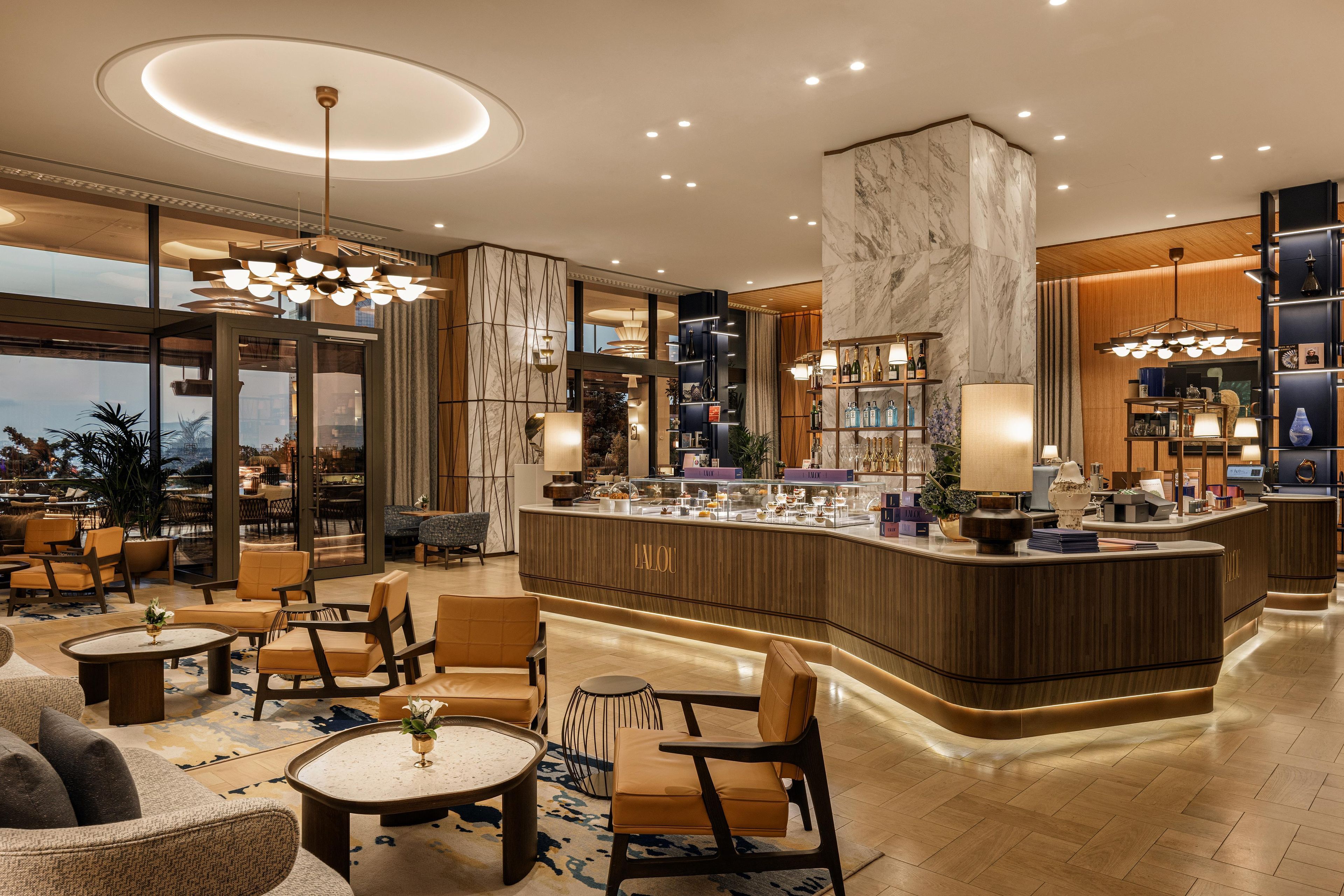 A modern hotel lobby with a central bar, seating areas, and a view through large windows.