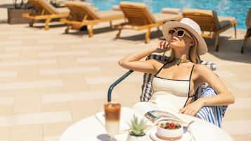 A woman relaxing by a poolside table with a drink, a small plant, and a plate of food.