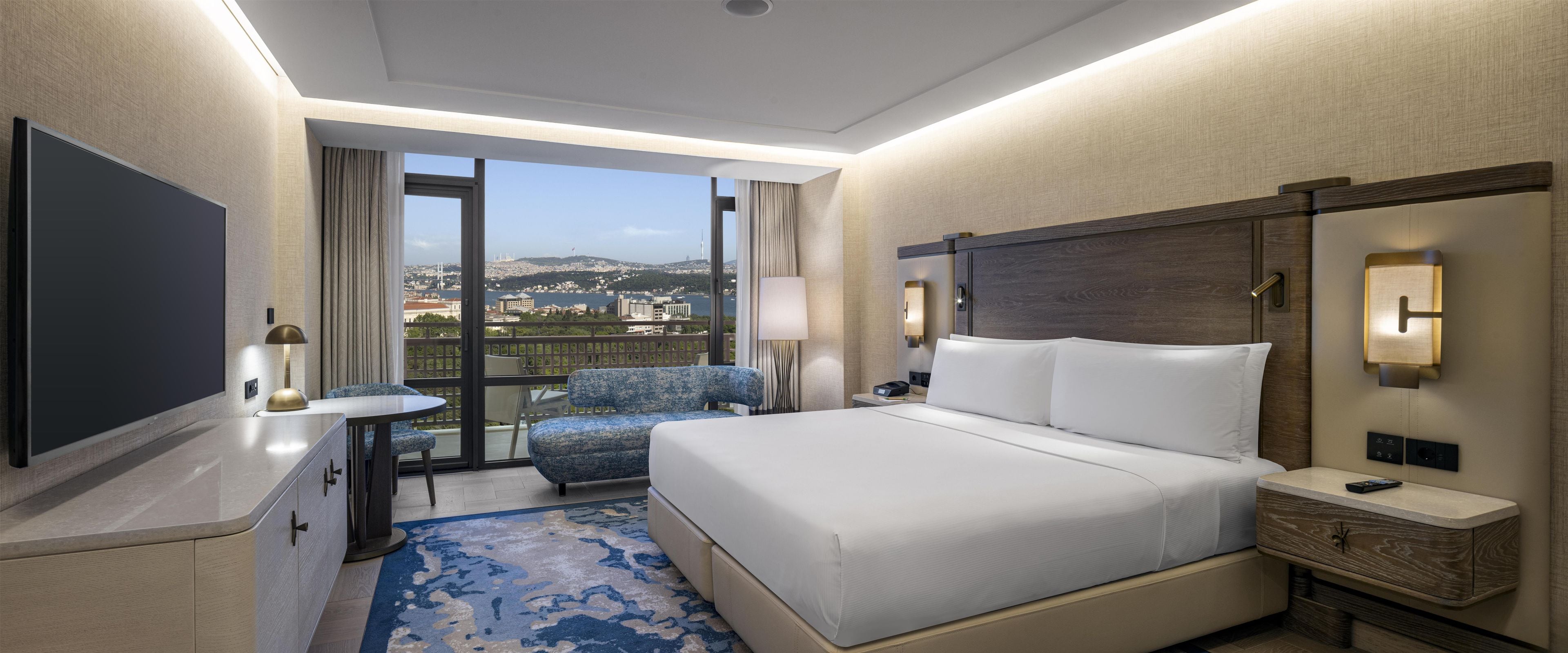 Habitación de hotel con una cama grande, un escritorio, una silla, un televisor y vistas a la ciudad.