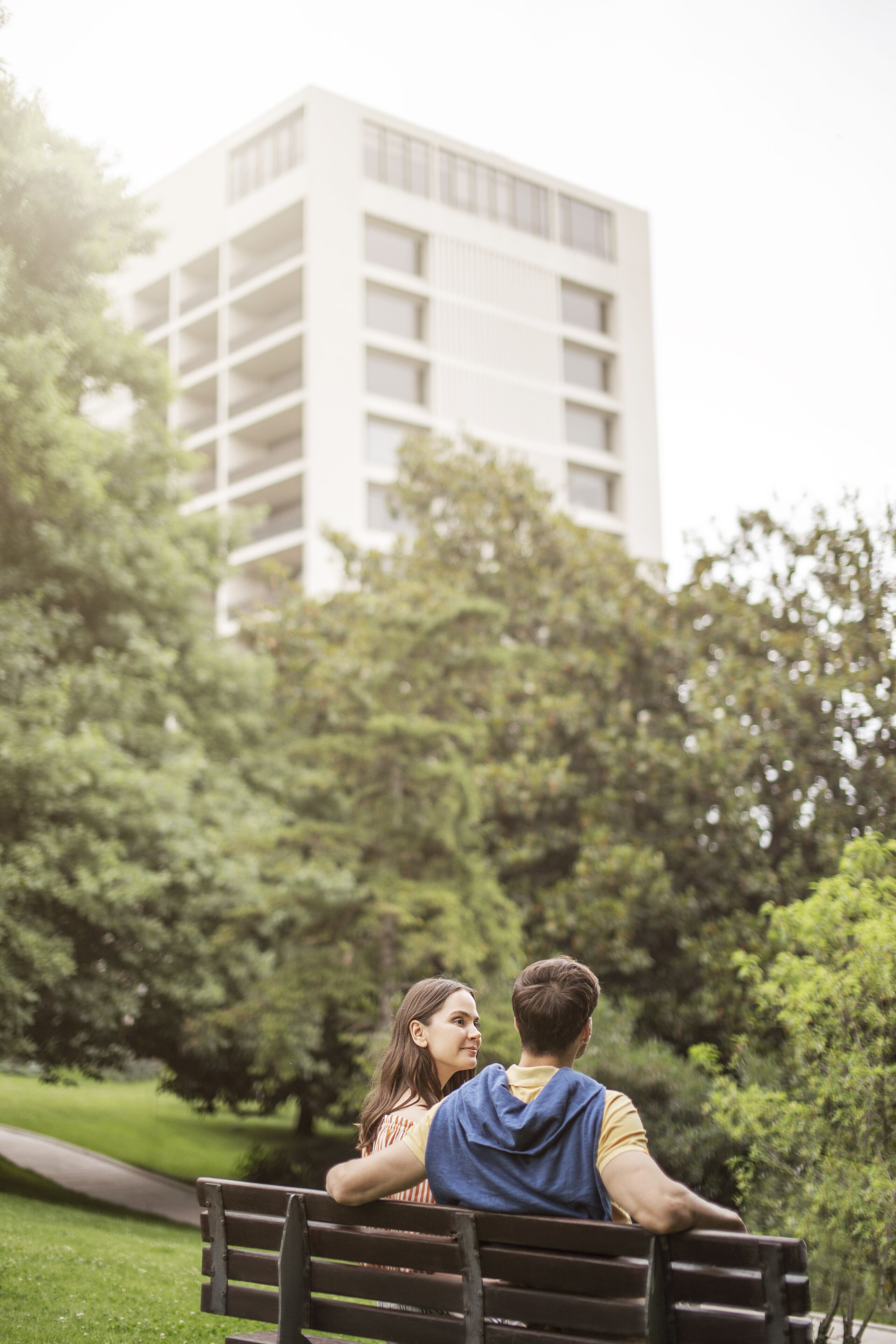 A couple sitting on a bench in a park with a modern building in the background.