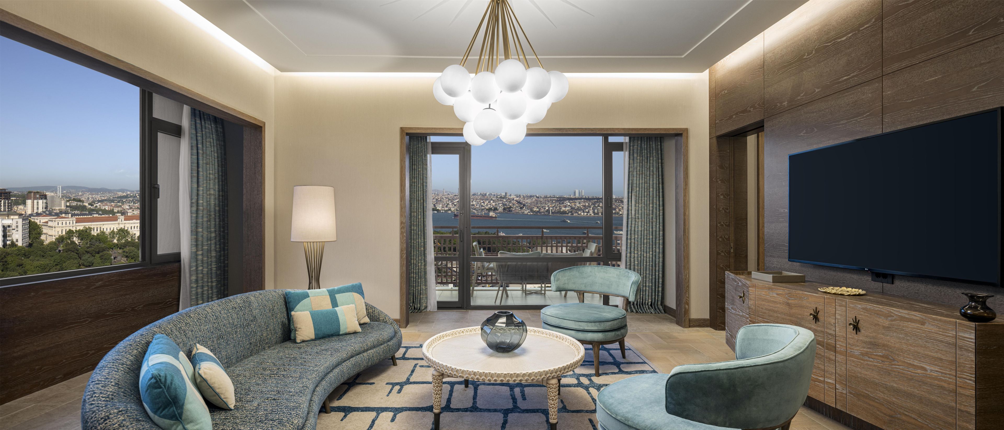 A modern living room with a sofa, armchair, and a round coffee table. There is a large TV on the wall and a view of a cityscape through the windows.