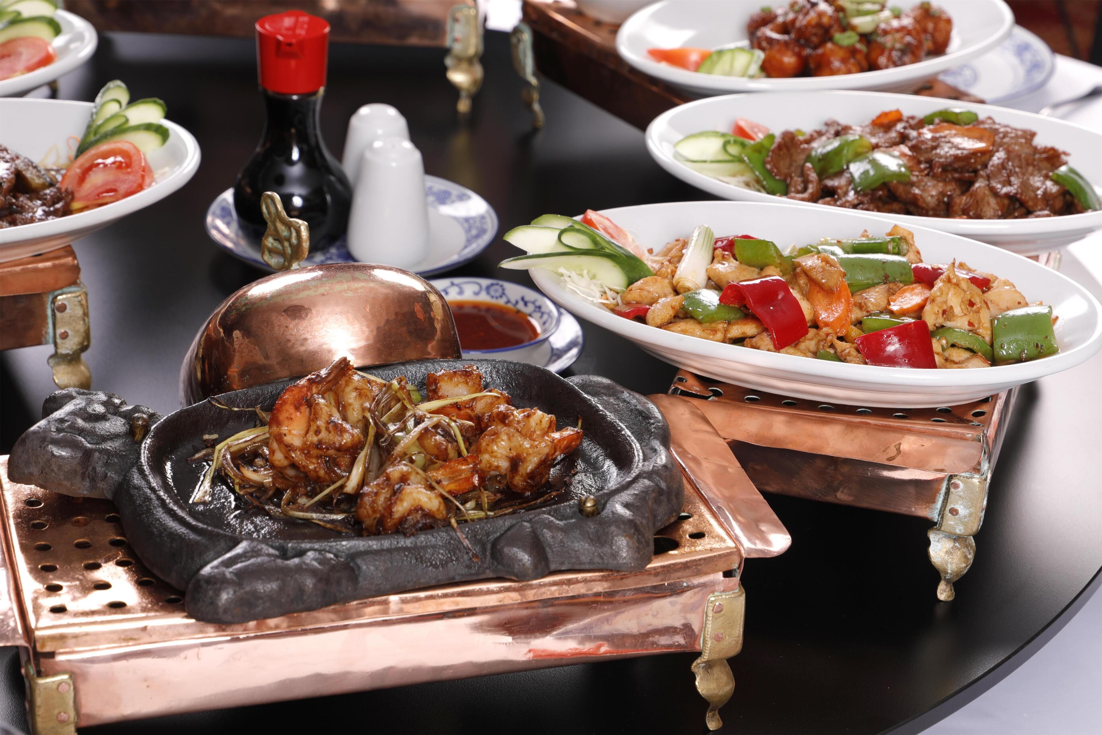 A table with various dishes, including a hot plate with food, a bottle of sauce, and several plates of vegetables and meat.