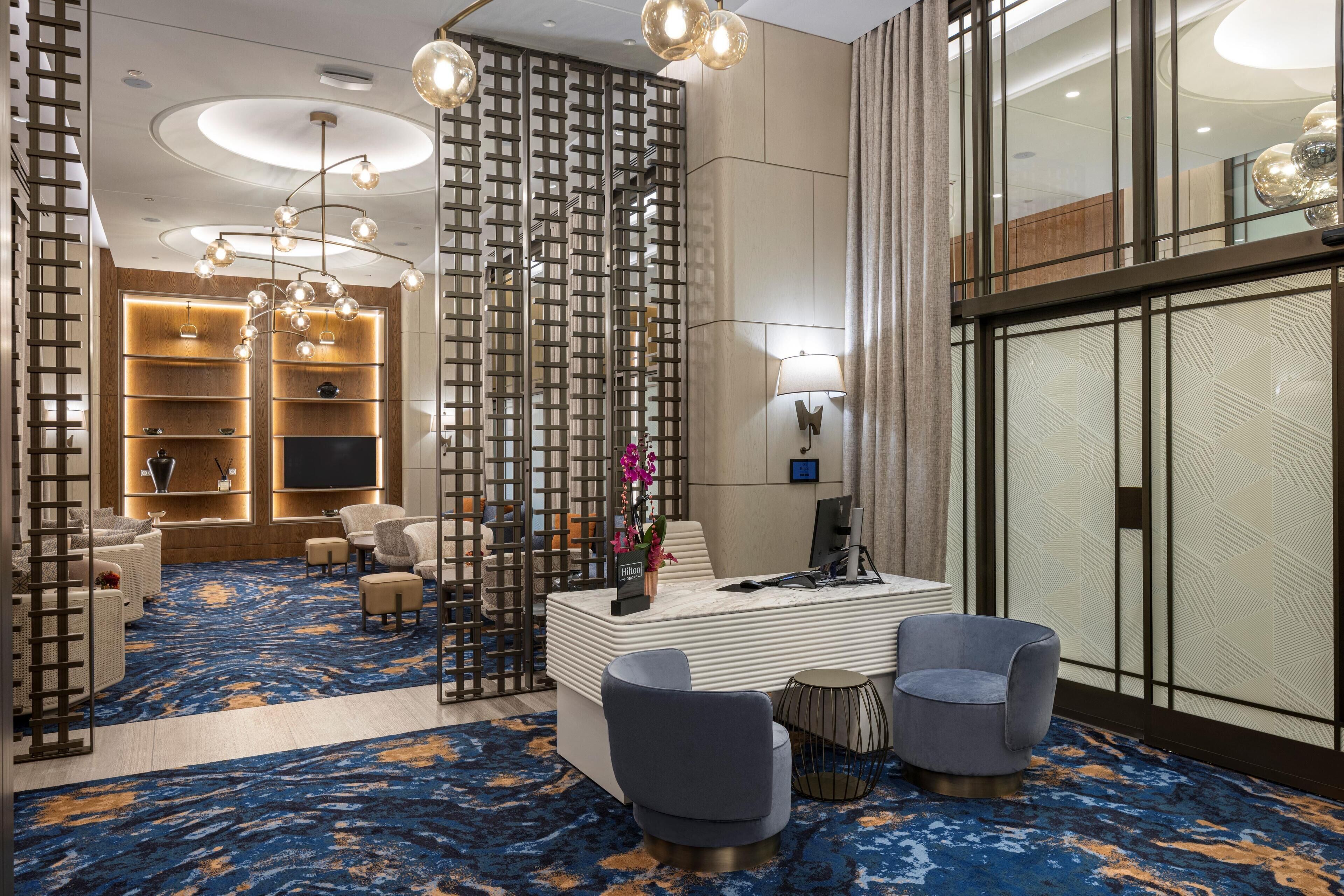 Un hall d’hôtel moderne avec un tapis à motifs bleus, un comptoir d’accueil, un espace salon et un mur décoratif orné d’un motif géométrique.