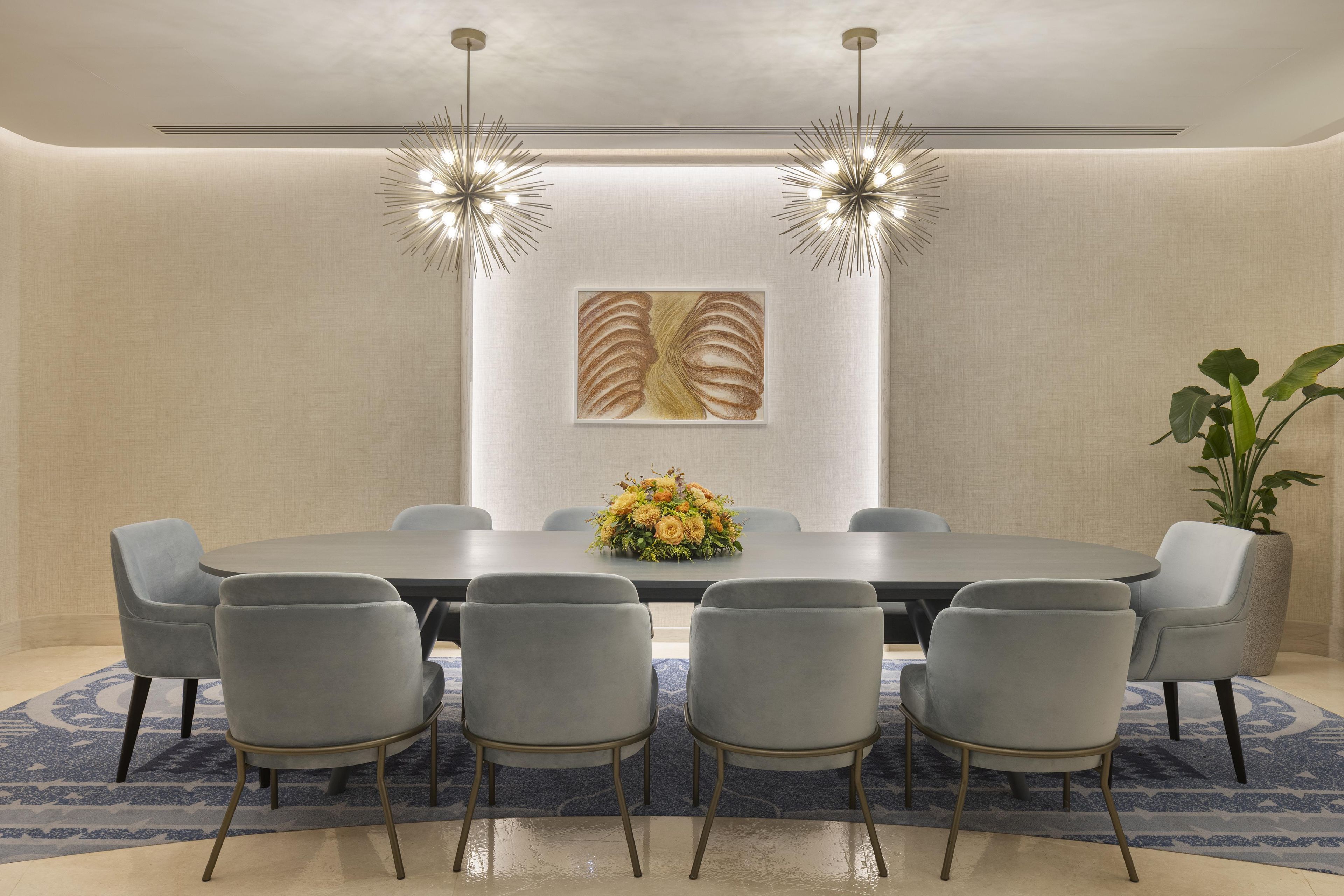 Une salle de conférence avec une longue table, six chaises, deux lampes suspendues, un tableau encadré et une plante en pot.