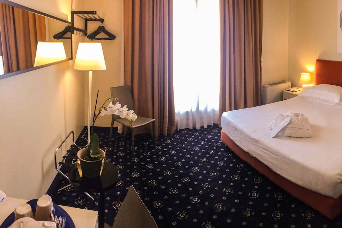 A hotel room with a large bed, two bedside tables with lamps, a chair, a desk with a lamp, a mirror, and a window with curtains.