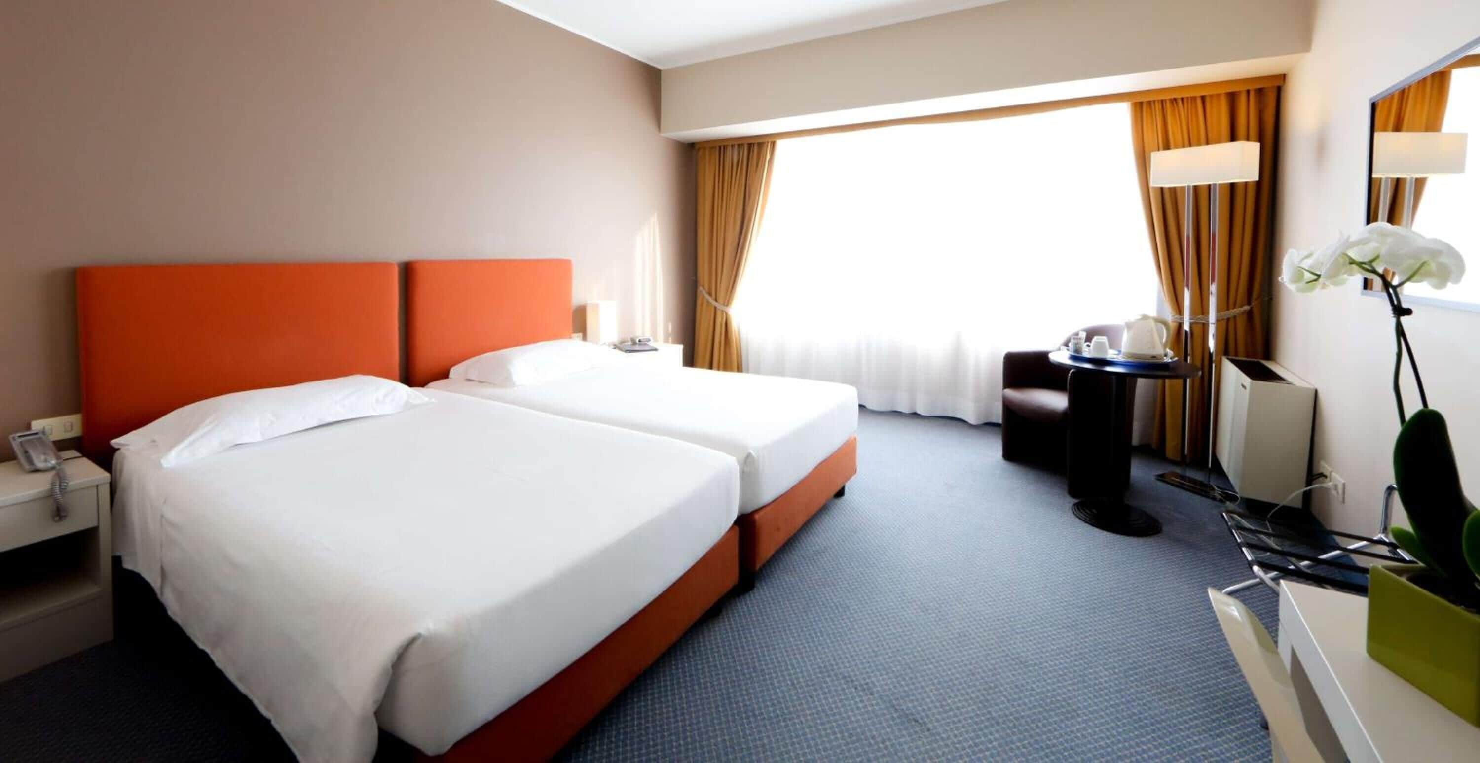 A hotel room with two beds, a desk, a chair, a lamp, and a window with curtains.