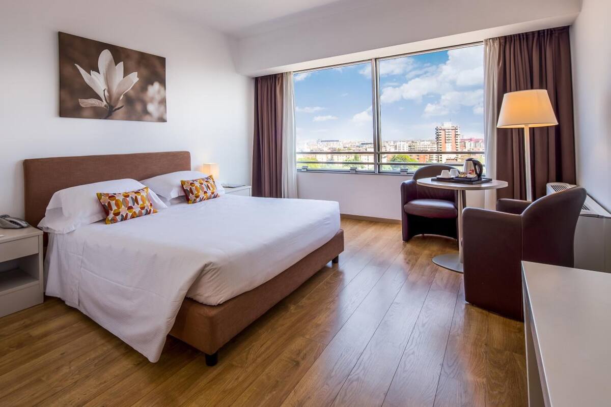 A hotel room with a large bed, a desk, a chair, a lamp, and a window with a city view.