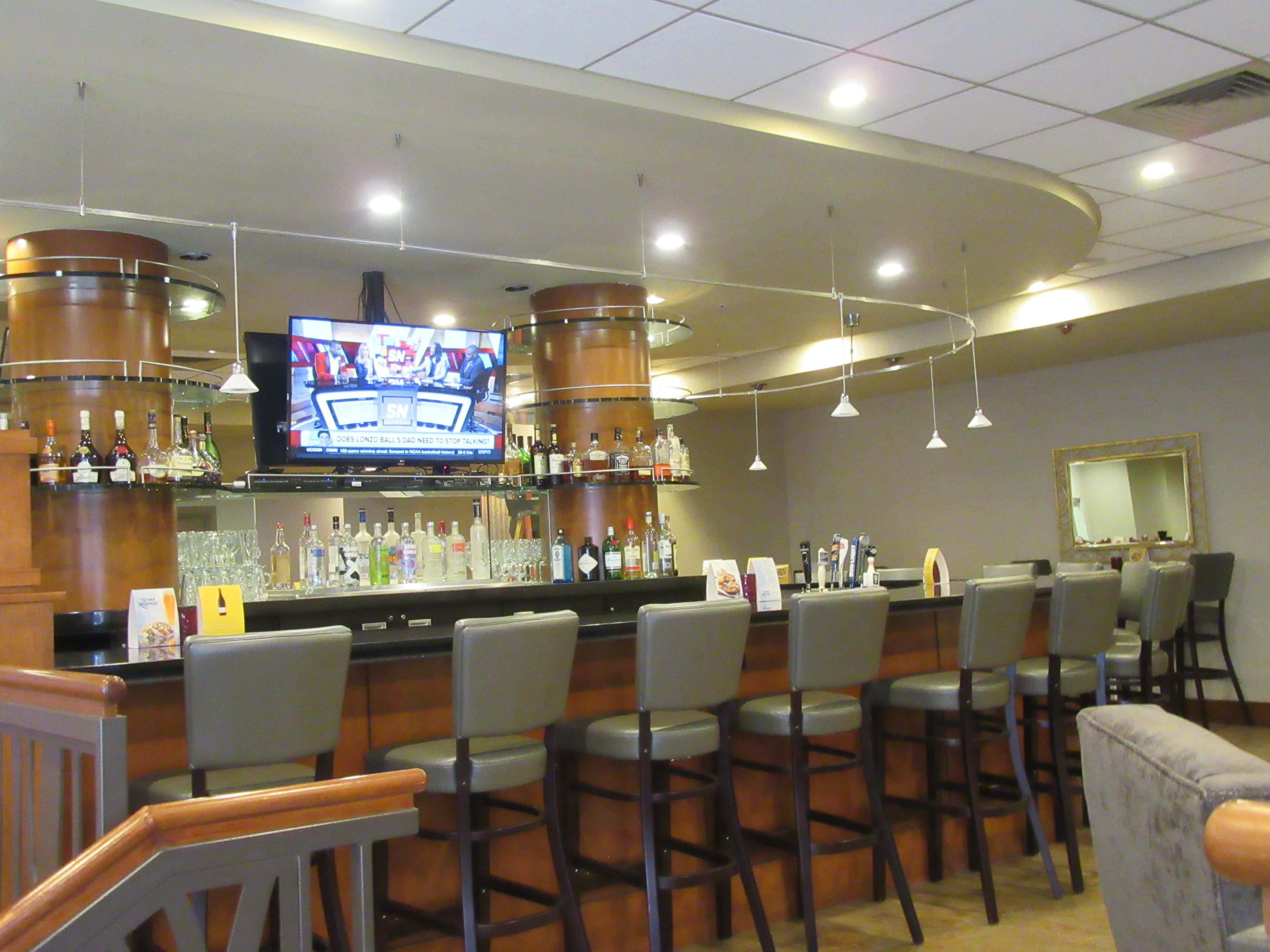 A bar area with multiple stools, a television mounted on the wall, and a variety of bottles and glasses on shelves.