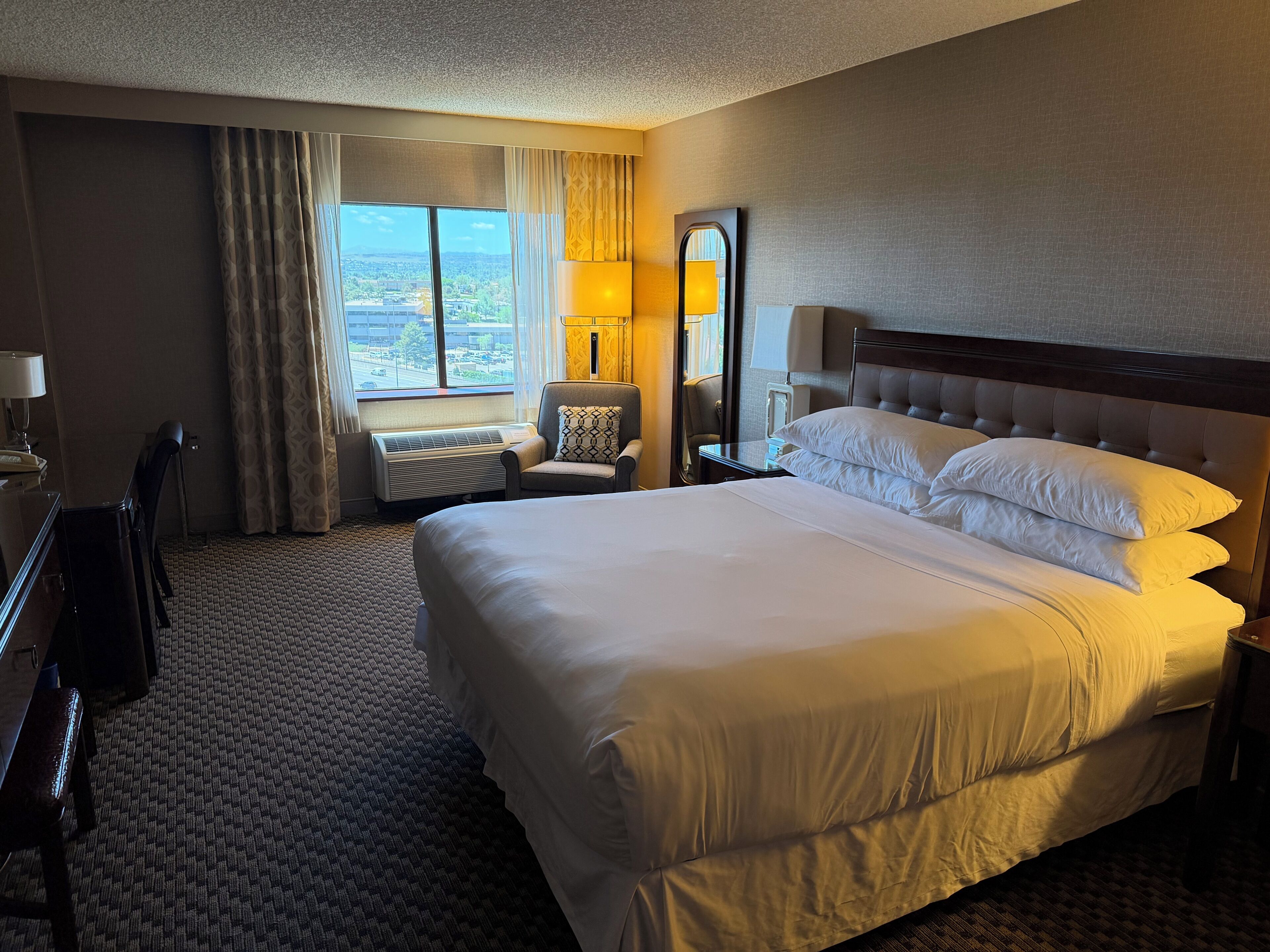 A hotel room with a large bed, a desk, a chair, a nightstand, a lamp, and a view of the city.