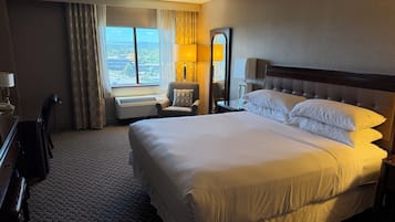 Ein Hotelzimmer mit einem großen Bett, einem Schreibtisch, einem Stuhl, einem Nachttisch, einer Lampe und Blick auf die Stadt.