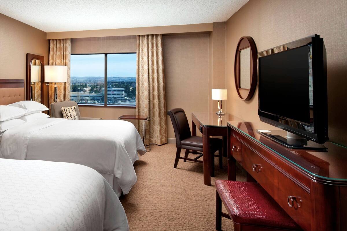 Een hotelkamer met twee bedden, een bureau met stoel, een televisie en een groot raam met uitzicht op de stad.