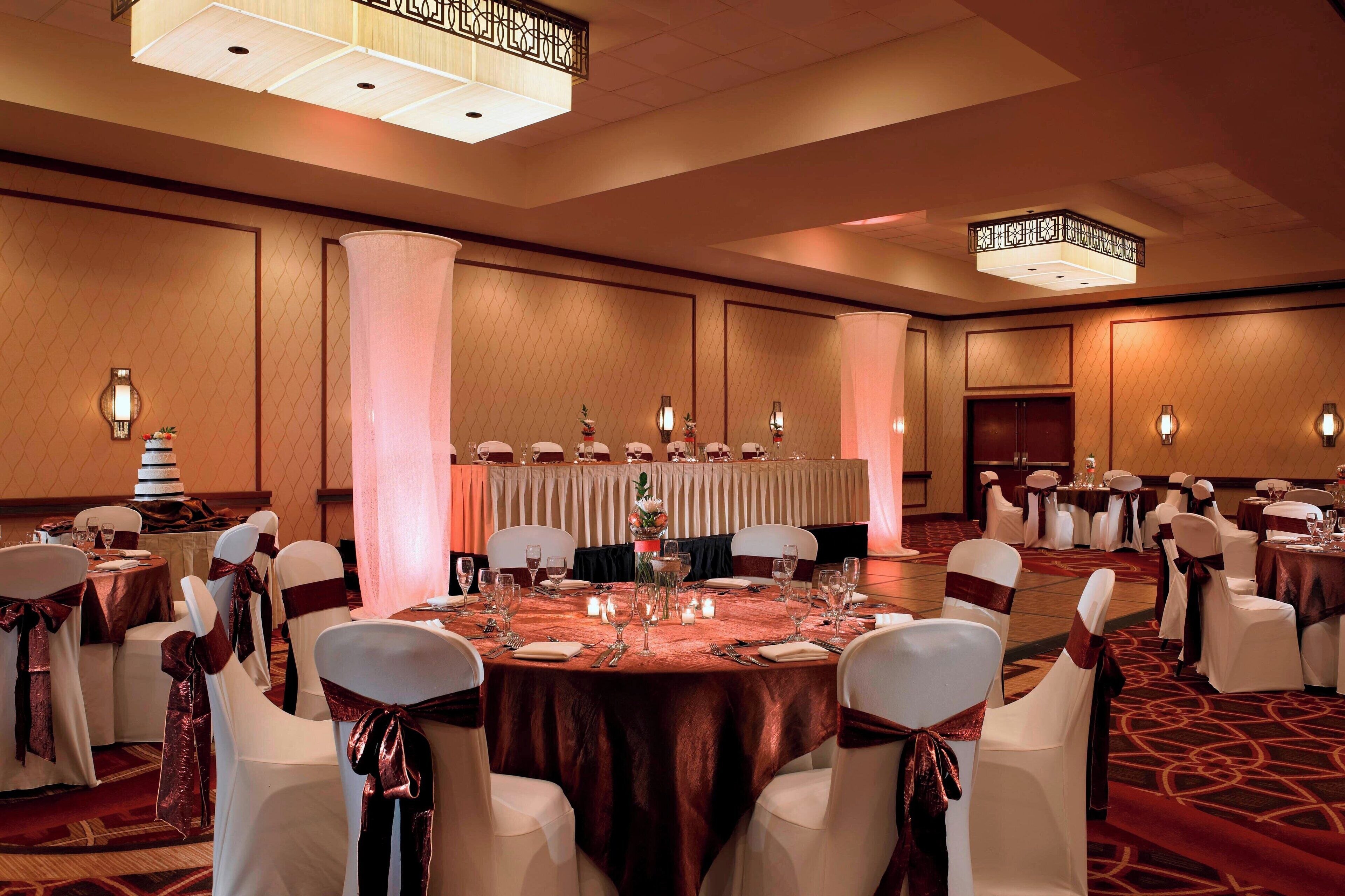 A banquet hall with round tables, white chairs with red sashes, and chandeliers.