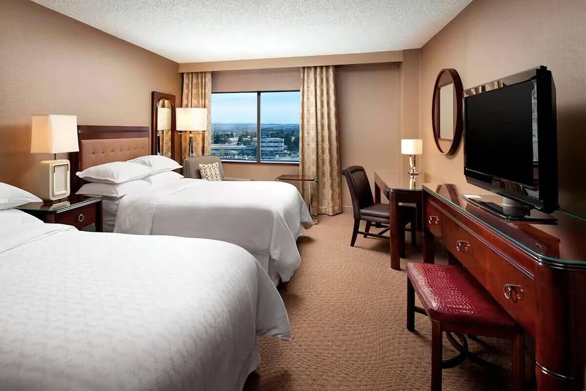 Een hotelkamer met twee bedden, een bureau, een stoel, een televisie en een groot raam met uitzicht op de stad.