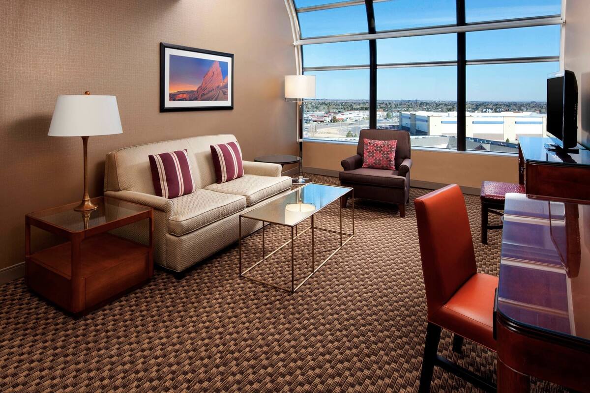 Een hotelkamer met een bank, fauteuil en uitzicht op de stad door grote ramen.