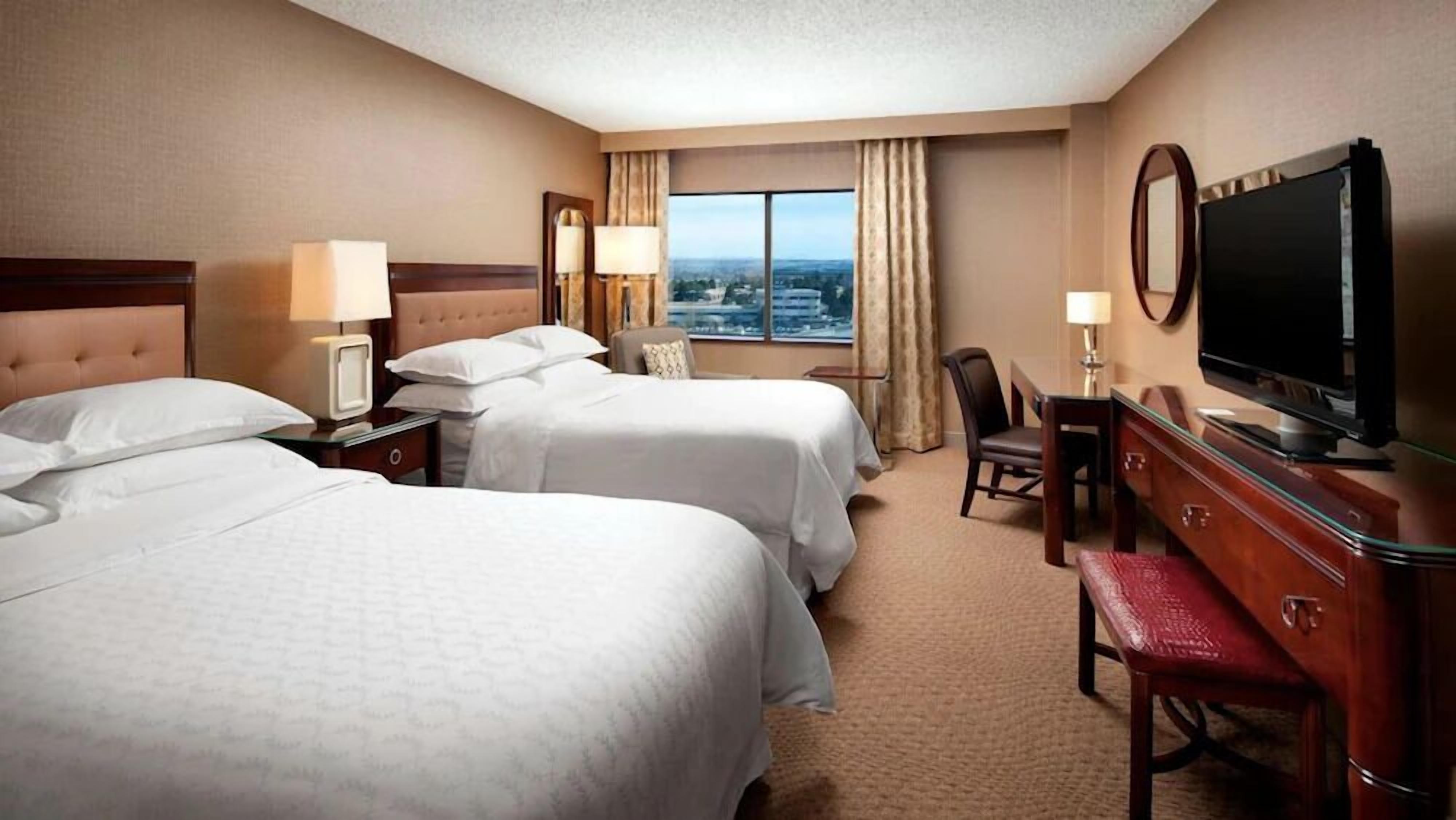 Ein Hotelzimmer mit zwei Betten, einem Fernseher, einem Schreibtisch und Blick auf die Stadt.