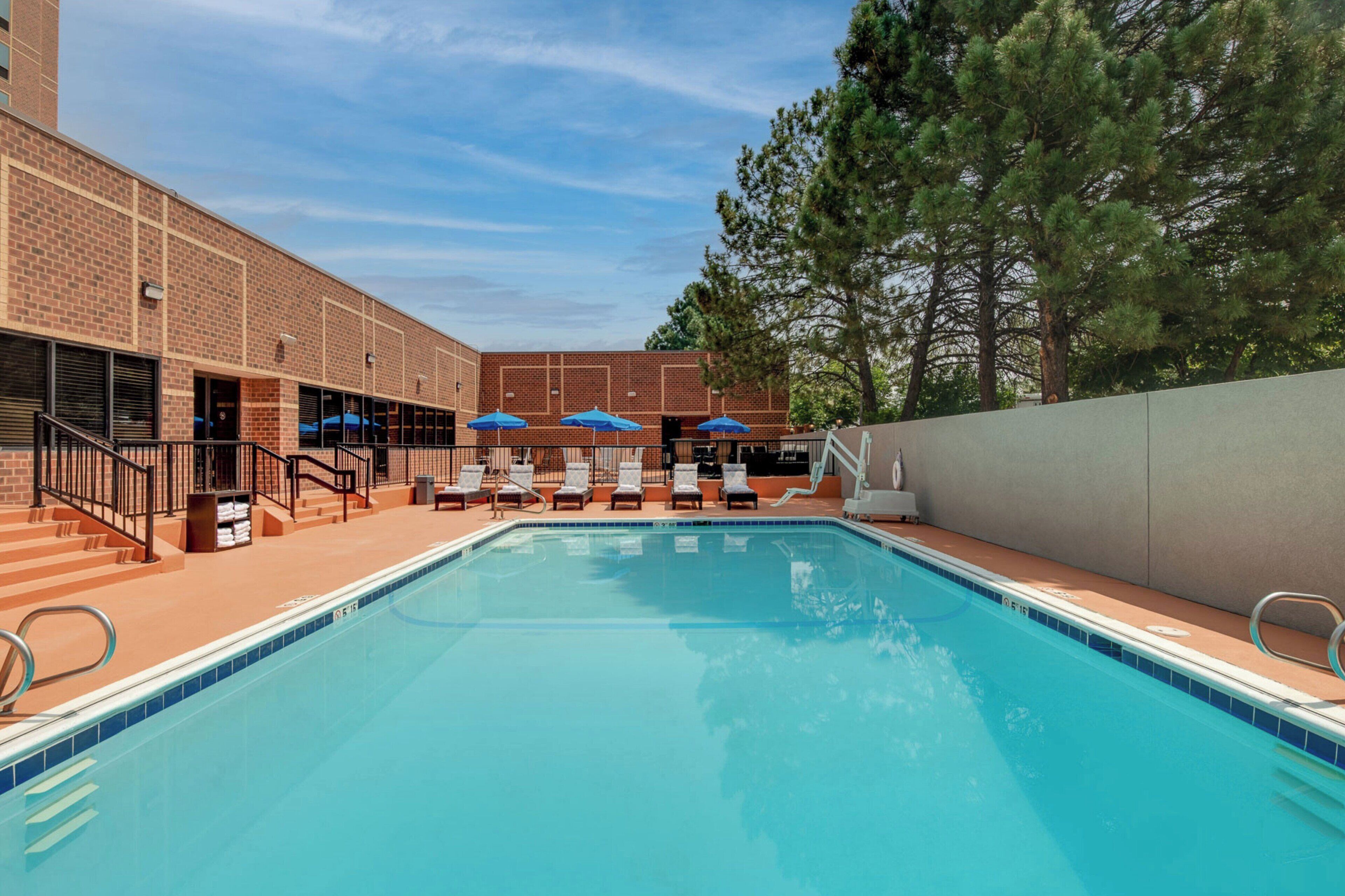A swimming pool with lounge chairs and umbrellas, surrounded by a brick building and trees.