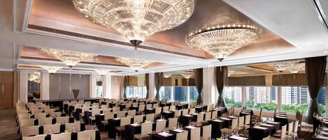 A conference room with rows of tables and chairs, large chandeliers, and a city view through the windows.