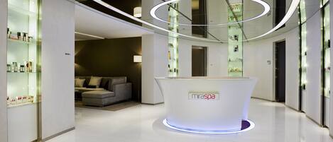Un vestíbulo moderno con una recepción que dice "MiraSpa", una vitrina con productos, una zona de estar con un sofá y un aplique de luz circular.