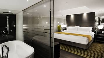 Habitación de hotel moderna con separador de vidrio que divide el baño y el dormitorio. El baño tiene una bañera grande y una zona de ducha. El dormitorio cuenta con una cama grande, una manta amarilla, una mesita de noche con lámpara y un espejo sobre la cama.