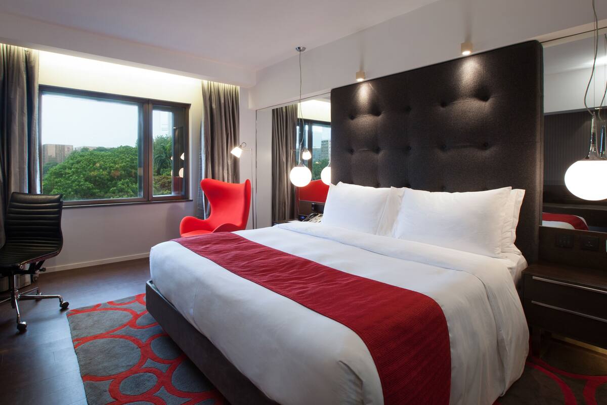 A modern hotel room with a large bed, a desk with a chair, and a view of greenery through the window.