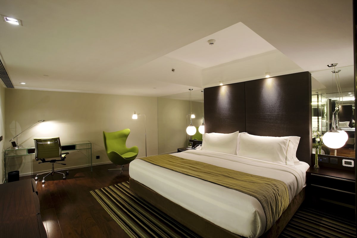 A modern hotel room with a large bed, a desk with a chair, a green chair, and a mirror.