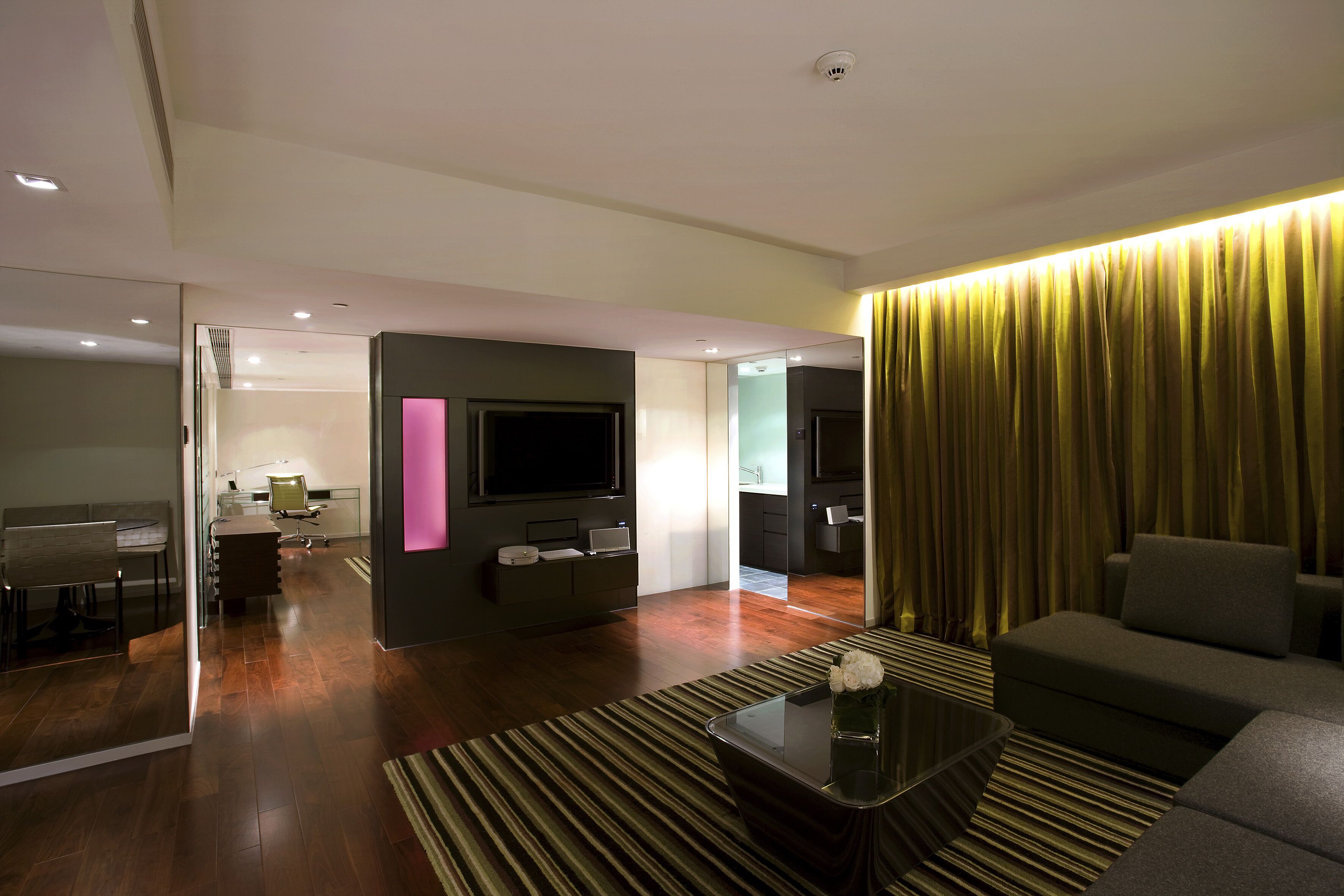 A modern living room with a sofa, a coffee table, and a TV. There is a kitchen area in the background with a dining table and chairs.