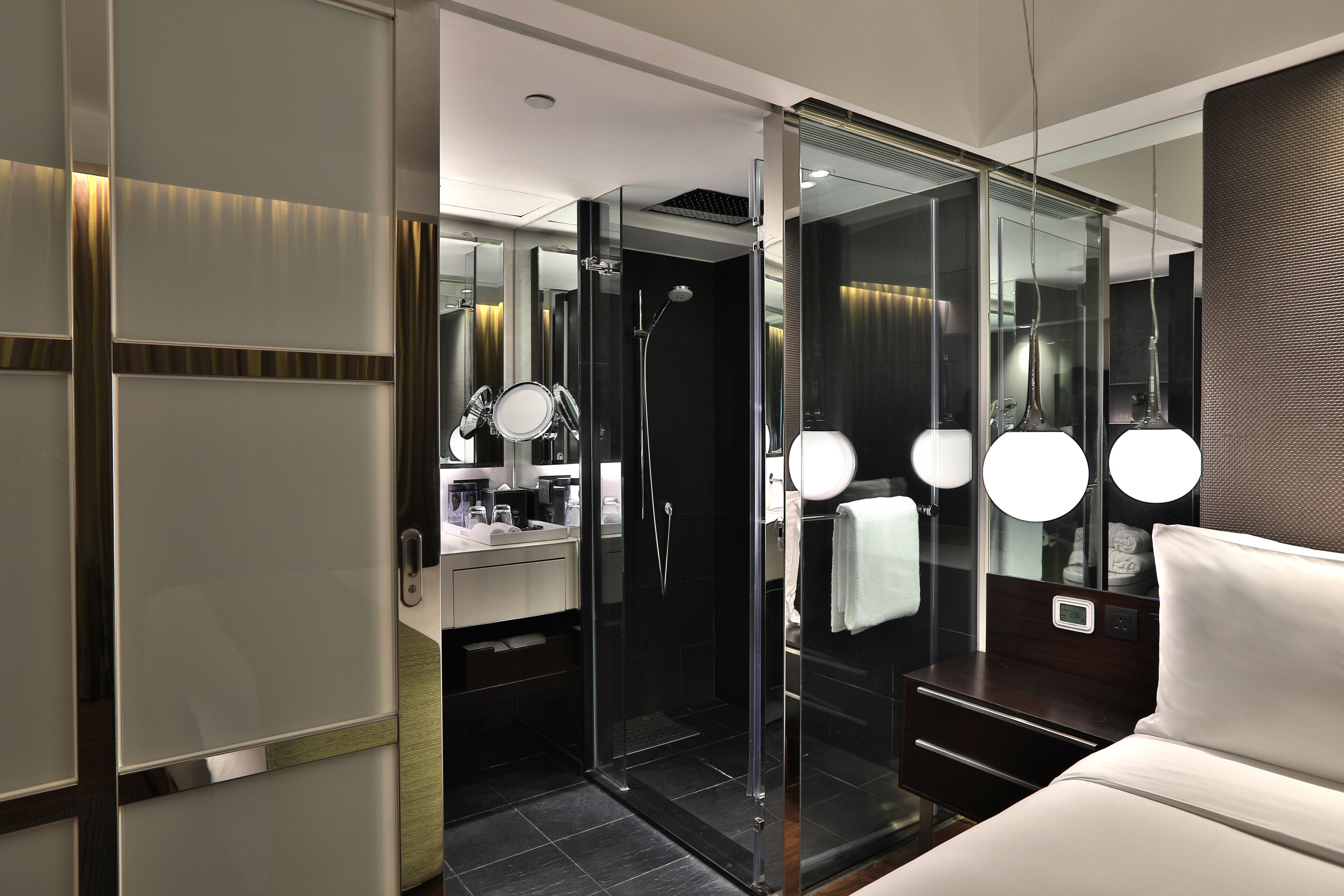 Ett modernt hotellbadrum med en duschavdelning med glasväggar, en tvättställsmöbel med spegel och en säng med sänggavel.