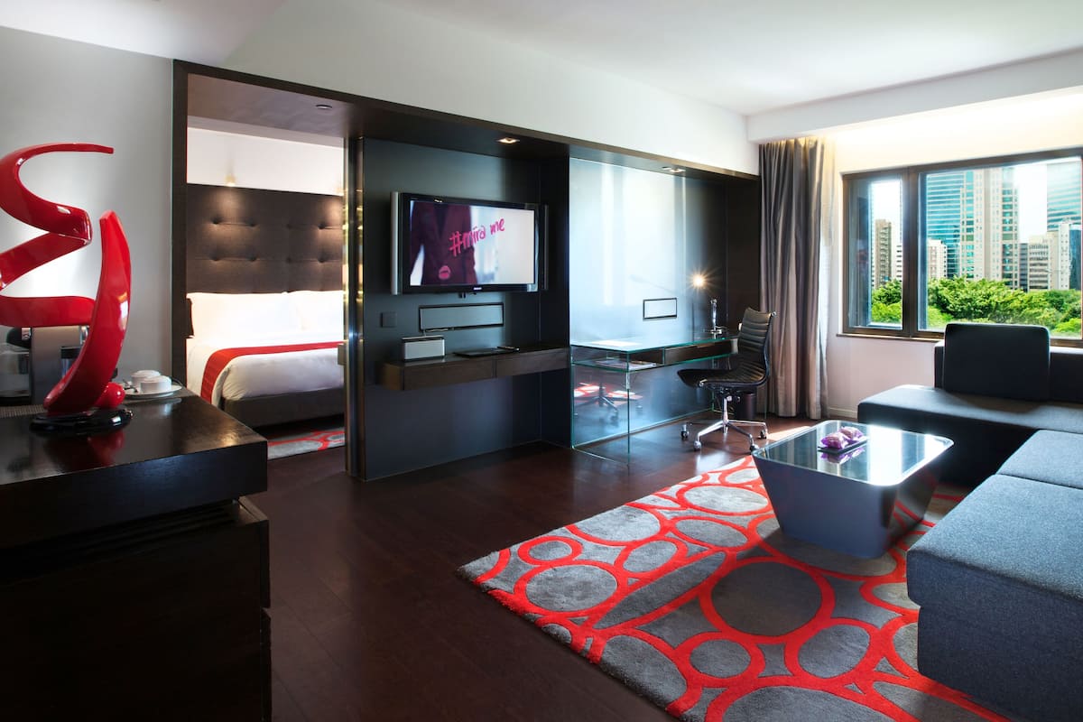 A modern hotel room with a bed, a TV, a desk, a sofa, and a large window with a city view.