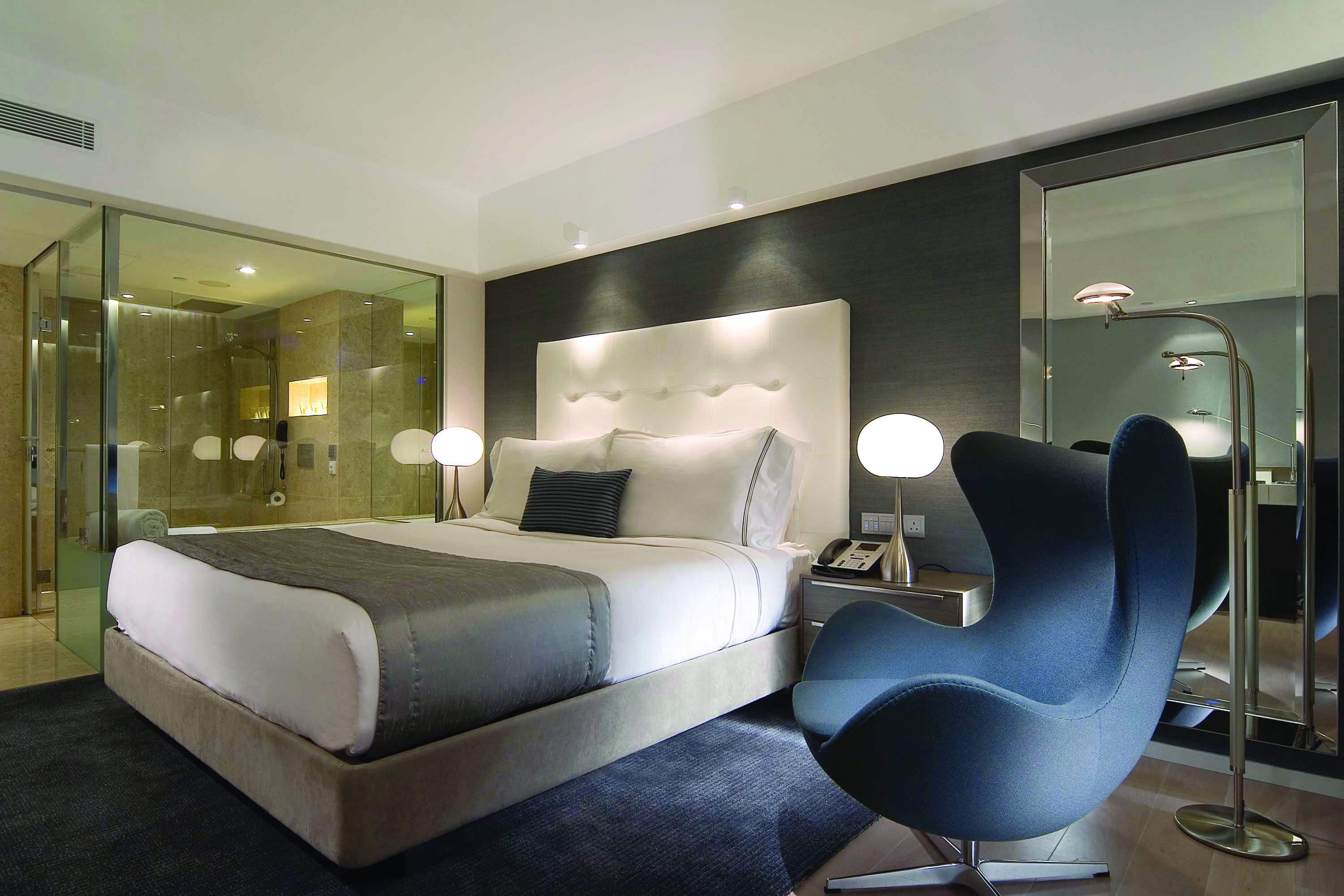 Een moderne hotelkamer met een groot bed, een nachtkastje met lamp, een blauwe stoel en een douchecabine van glas.