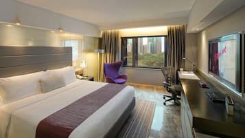 A hotel room with a large bed, a desk with a computer, a purple chair, and a view of the city through the window.