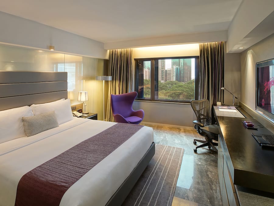 A hotel room with a large bed, a desk with a computer, a purple chair, and a view of the city through the window.