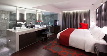 Una habitación de hotel moderna con una cama grande, un escritorio separado con una silla, un espejo grande y un televisor.