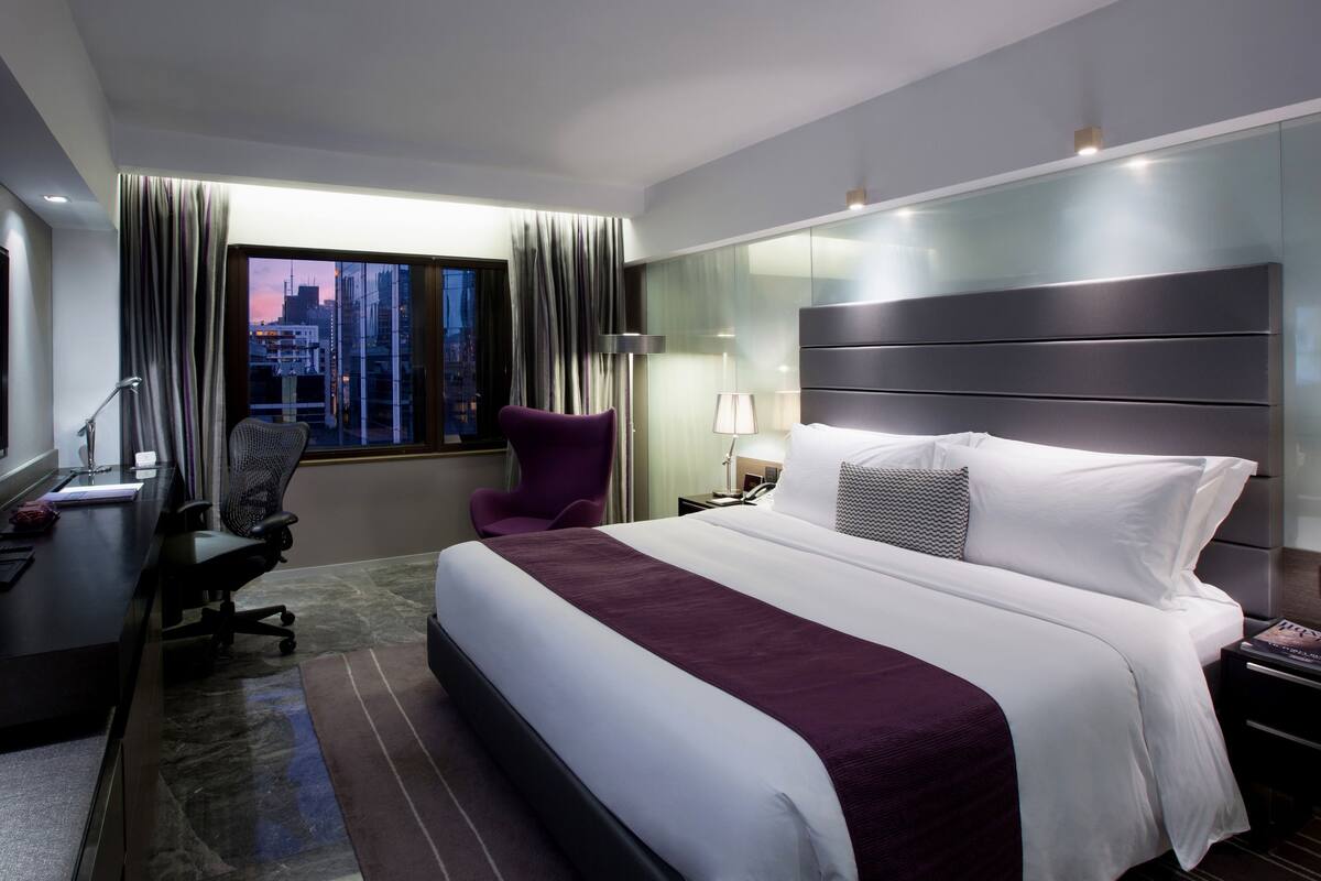 A modern hotel room with a large bed, a desk with a computer, a chair, and a TV.